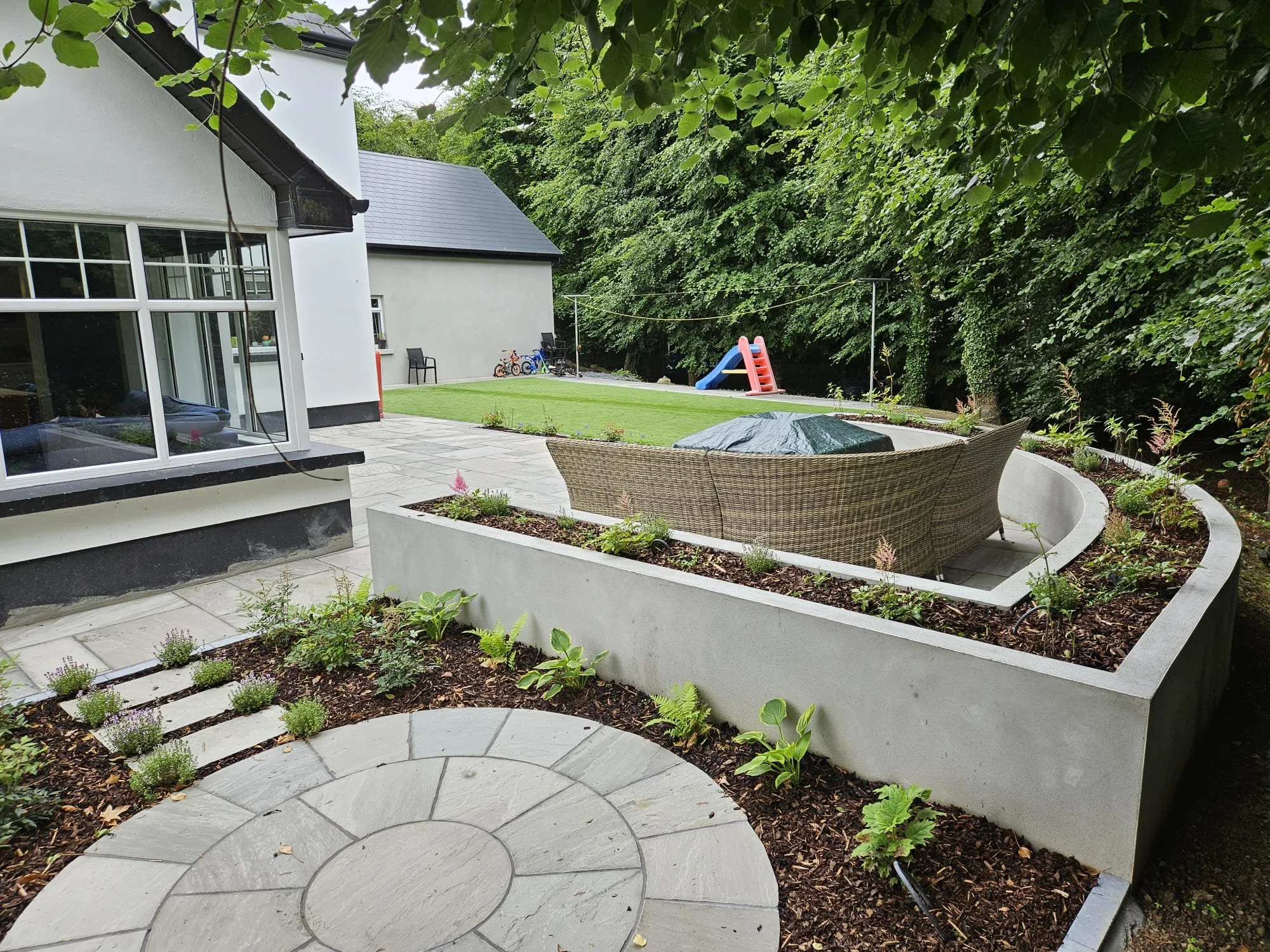 View of backyard patio with round stone pathway, garden beds with plants, wicker outdoor sofa with cover, and a well-maintained lawn with children's slide and bicycles in the background, surrounded by trees.