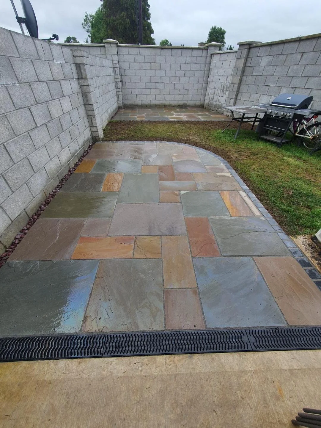 Backyard patio with multicolored stone tiles, grey brick wall, grassy area, grill, and a bike.