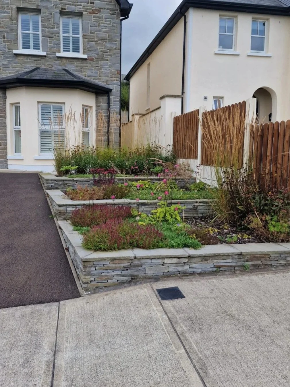 A landscaped front yard with a tiered flower bed made of stacked stone, featuring various plants and pink flowers, located between two houses with a paved driveway on the left.