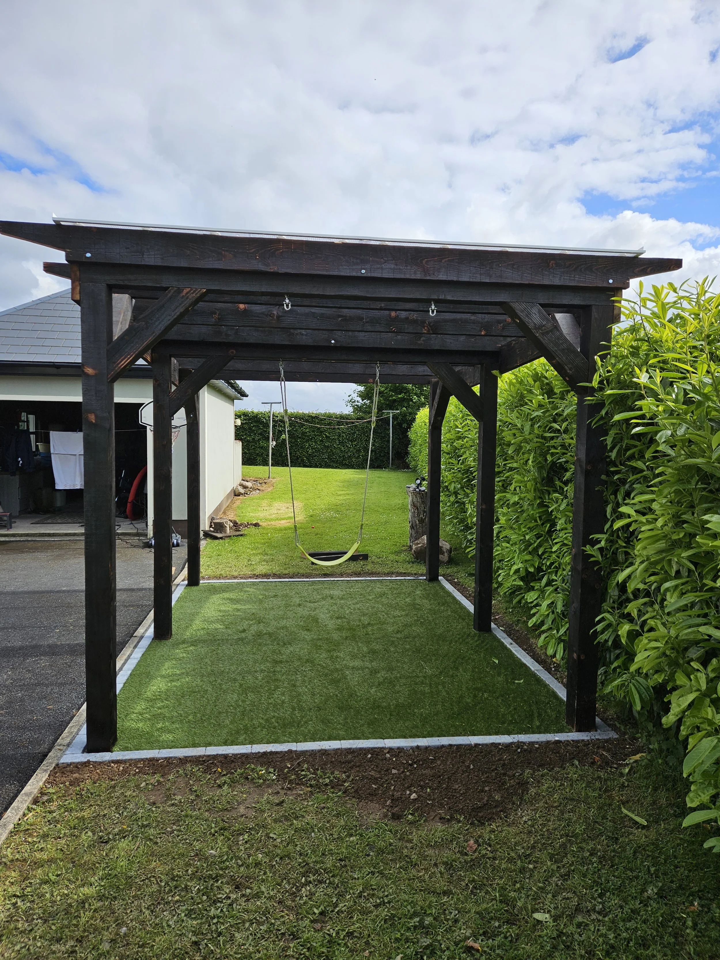 A backyard wooden gazebo with a roof, two swings hanging from the ceiling, a grassy lawn beneath, and green hedges on the right side. A neighboring house is visible on the left, with a basketball hoop mounted on the wall.