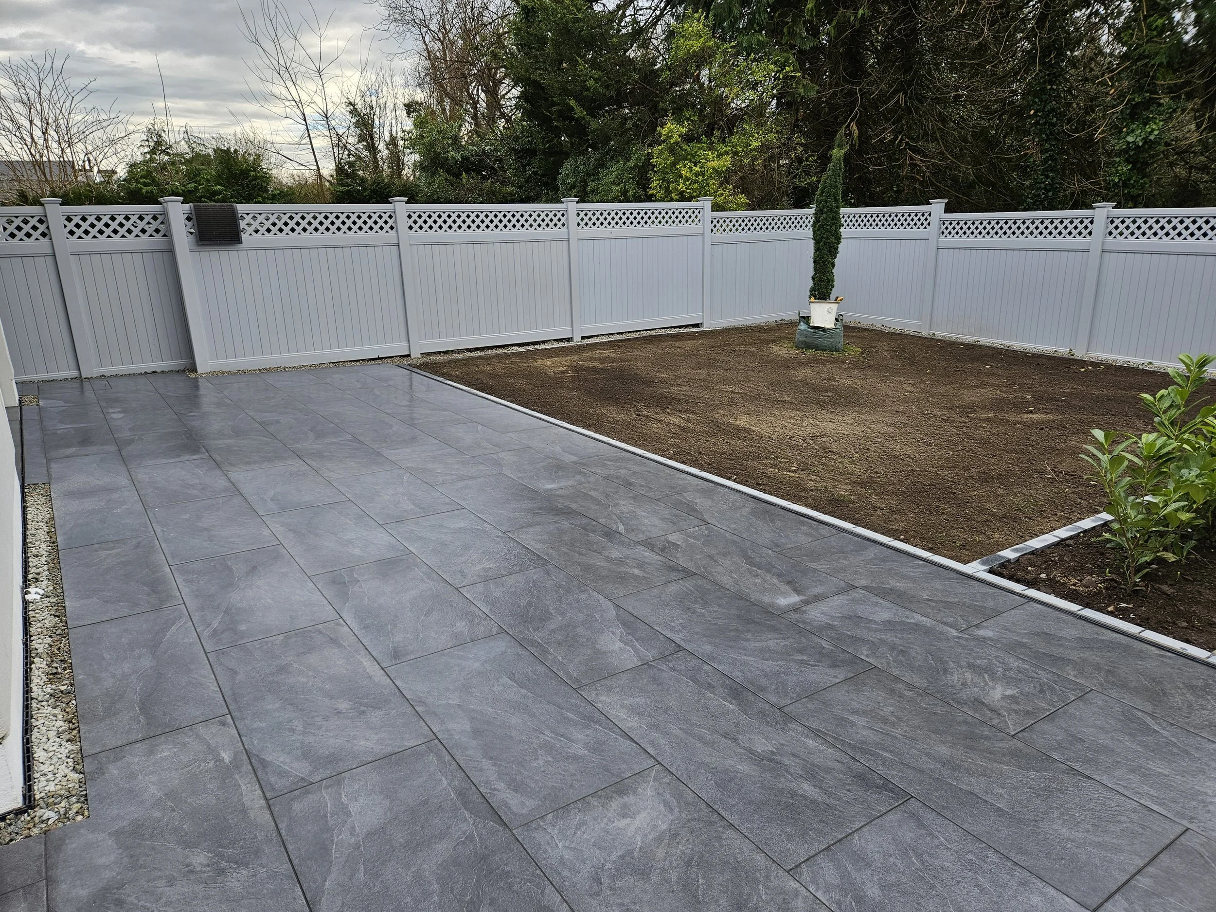A backyard scene showing a newly installed gray tiled patio area, a small garden bed with some plants, a white fence surrounding the yard, and a small potted tree.