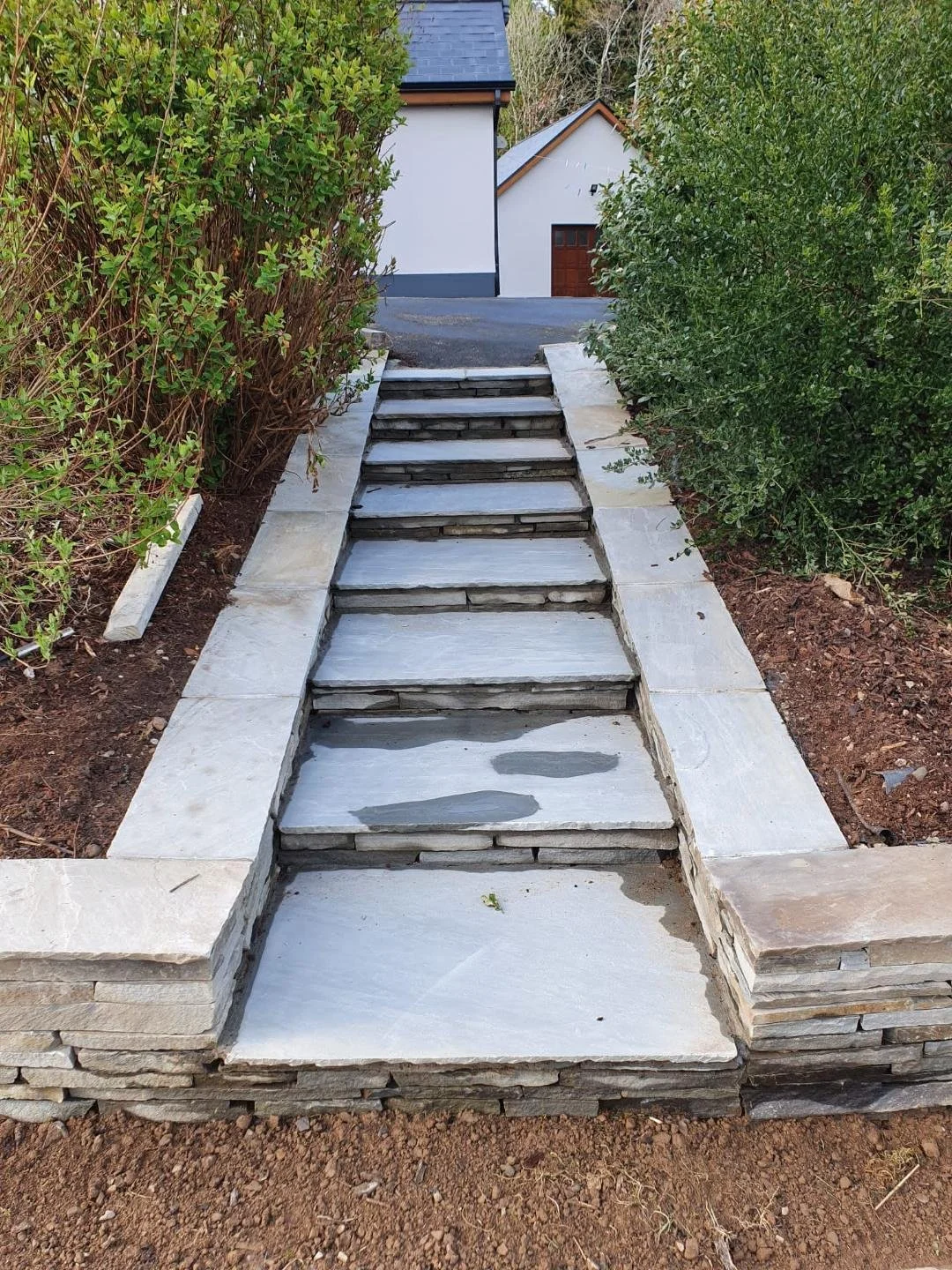 Stone stairway with wide white stone steps flanked by green bushes leading up to a paved driveway with a house in the background.