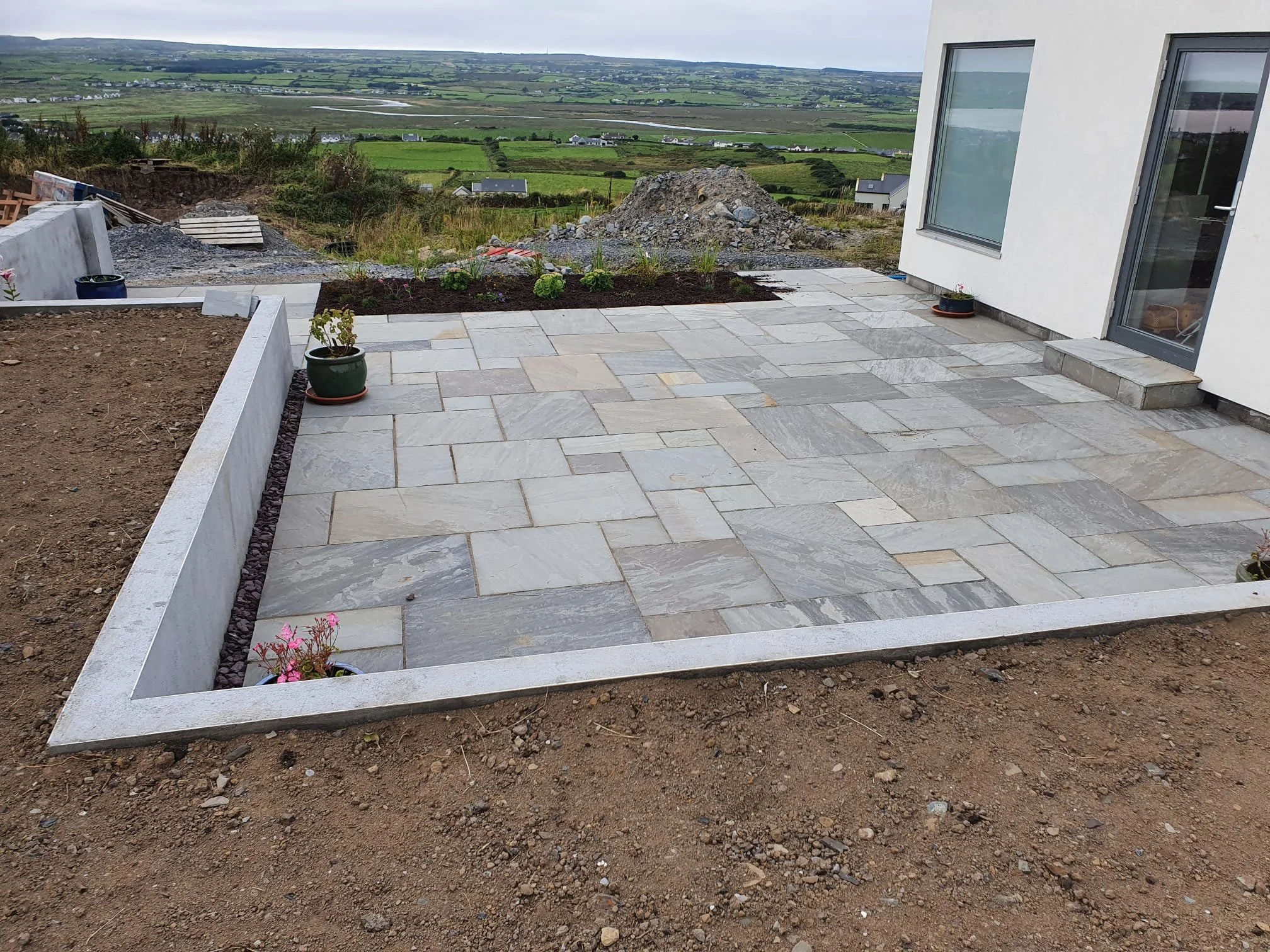 A patio with stone tiles next to a white house with a glass door, surrounded by a garden with potted plants and a view of rolling green hills in the background.