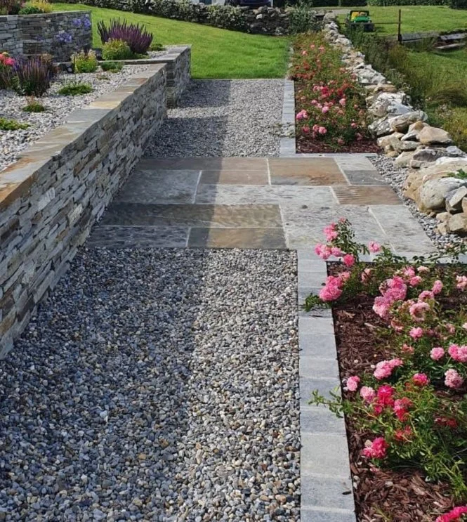 A landscaped garden pathway with gray stone pavers and gravel, bordered by flower beds with pink roses and a stone retaining wall on the left.