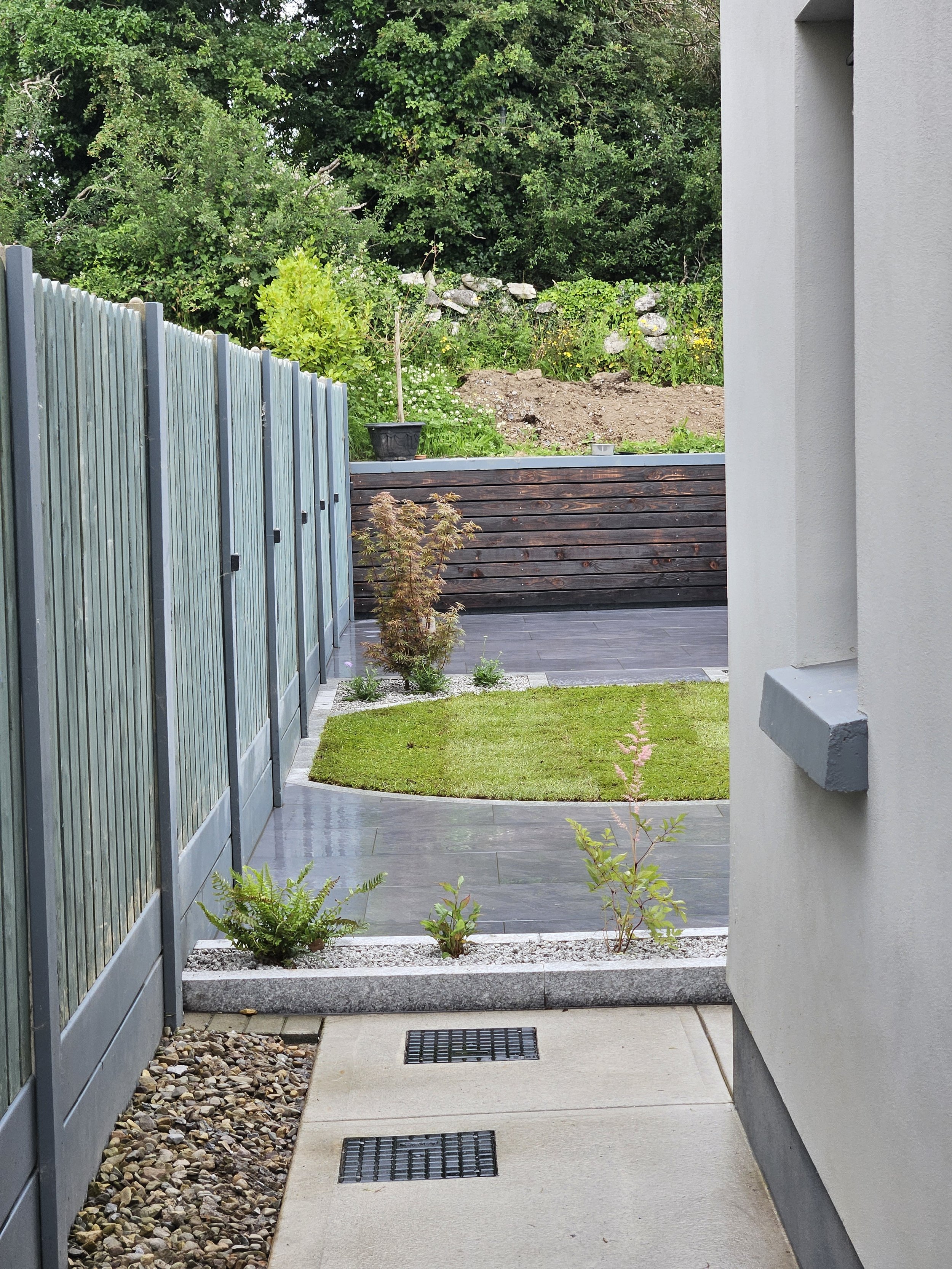 View of a modern backyard garden with a small patch of grass, young plants, a paved pathway, a wooden fence on the left, and a raised wooden deck at the back, with a background of trees and a stone wall.
