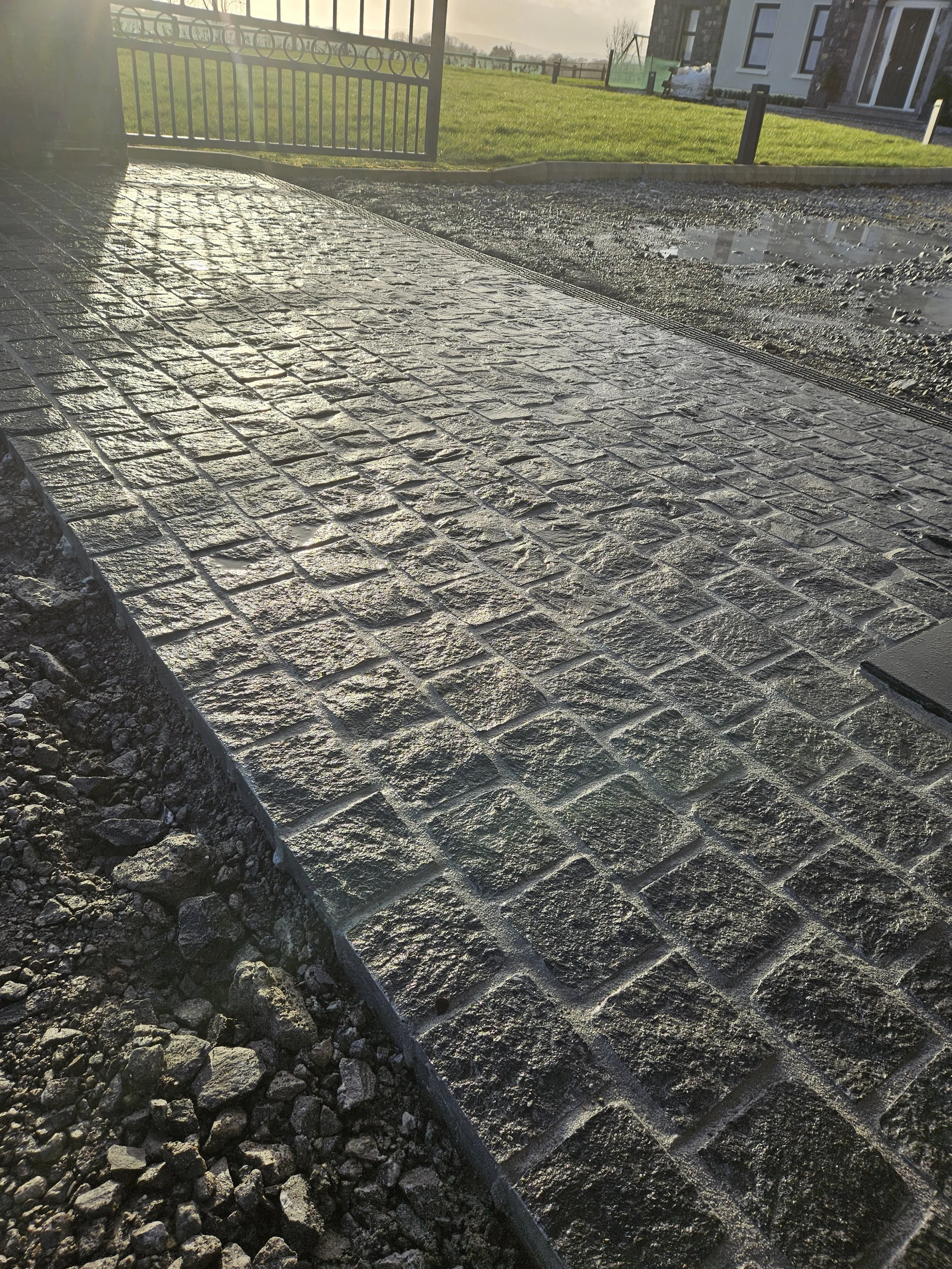 A newly paved brick walkway with textured bricks, with a section of gravel and dirt on the side, and a grassy area with a black metal fence and a house in the background.