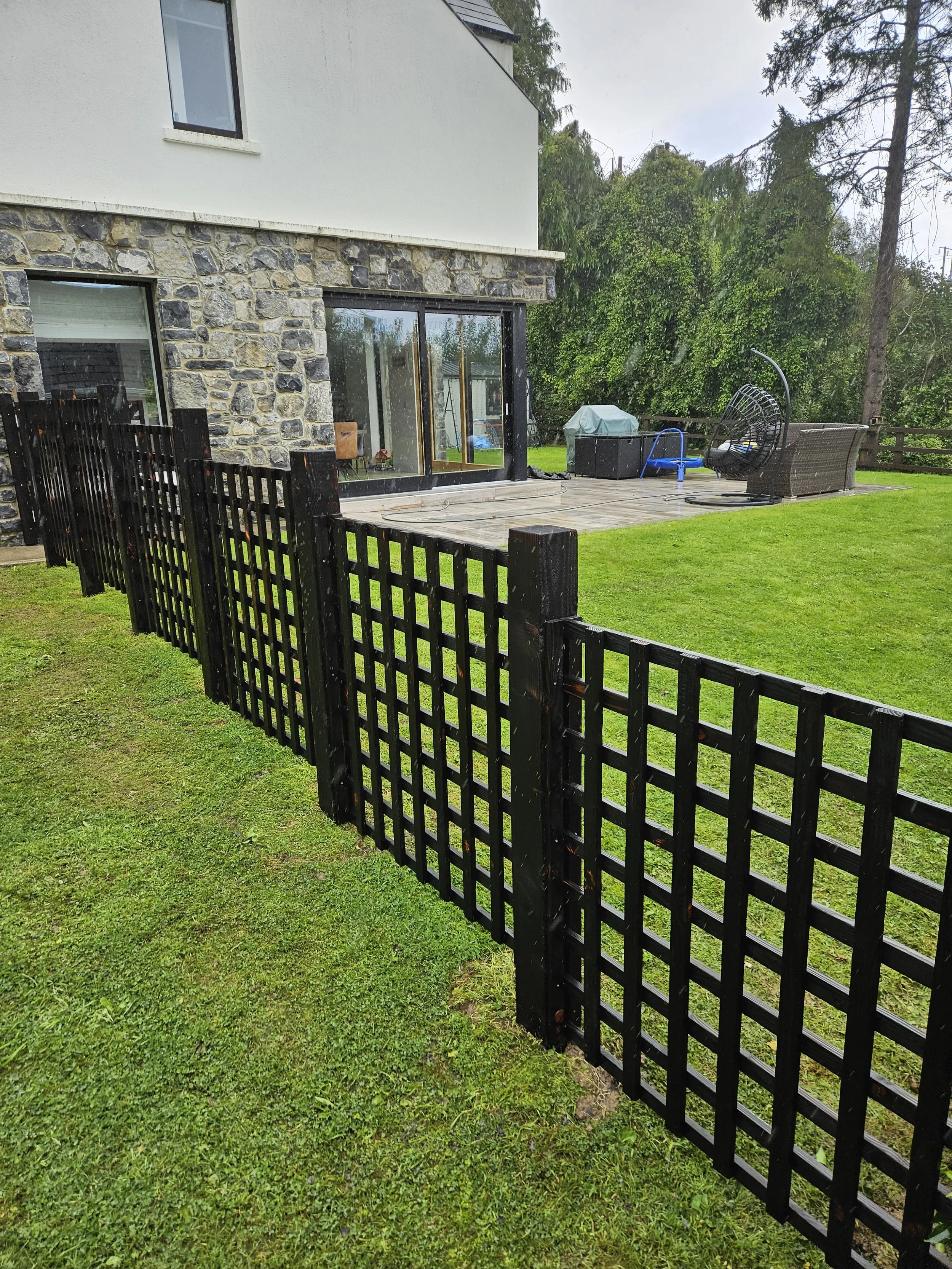 Black wooden fence surrounding a lush green backyard with a patio, outdoor furniture, and a garden