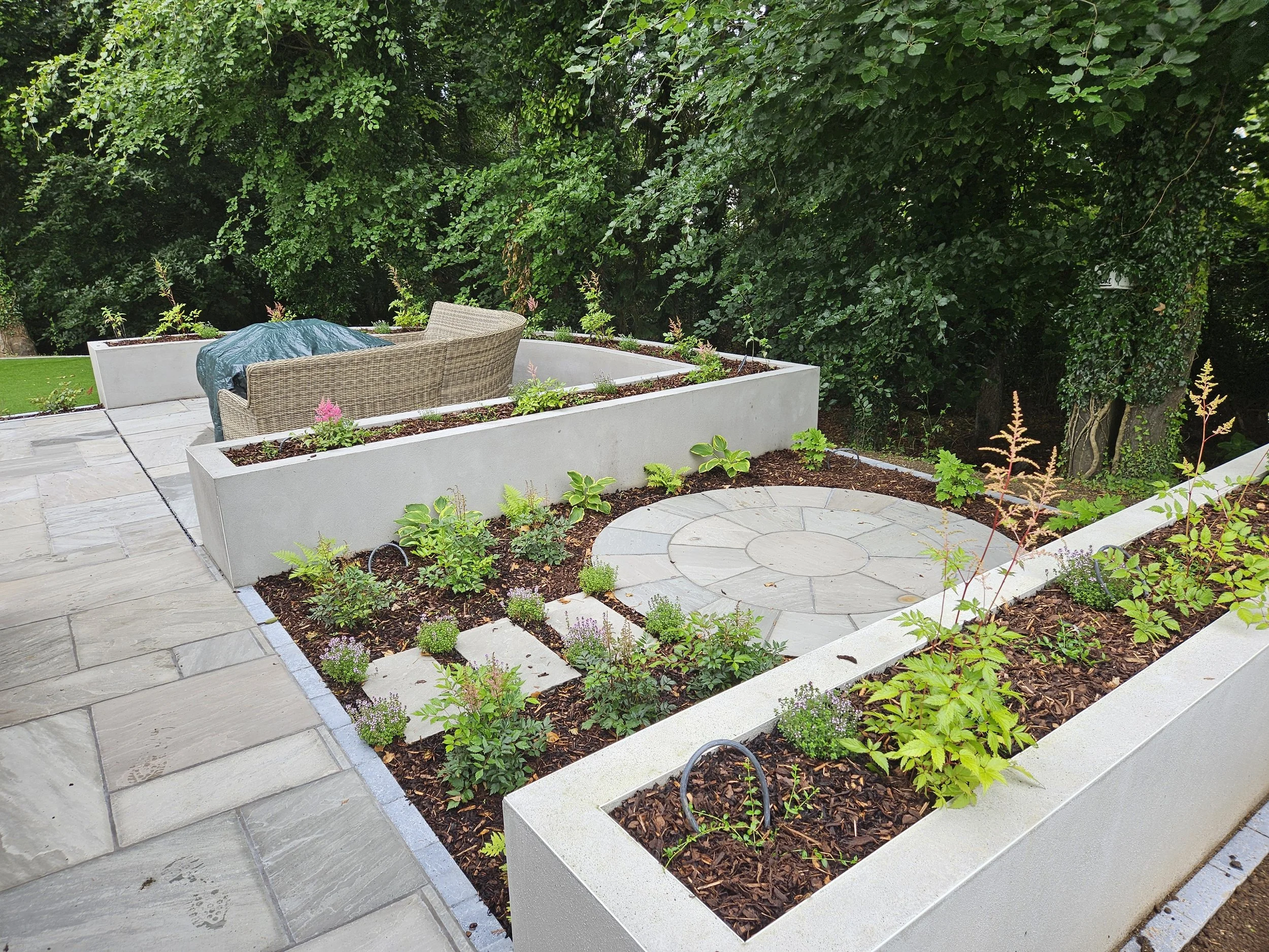A modern outdoor patio with paved flooring, white raised planters with green plants, and flowerbeds. One flowerbed contains a circular paved area with a garden sofa and a table covered with a teal cloth, surrounded by lush green trees and foliage.