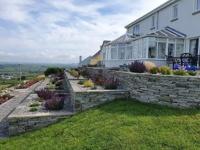 A house with a white exterior and a sunroom, overlooking a terraced garden with stone walls, purple and green plants, and a green lawn.