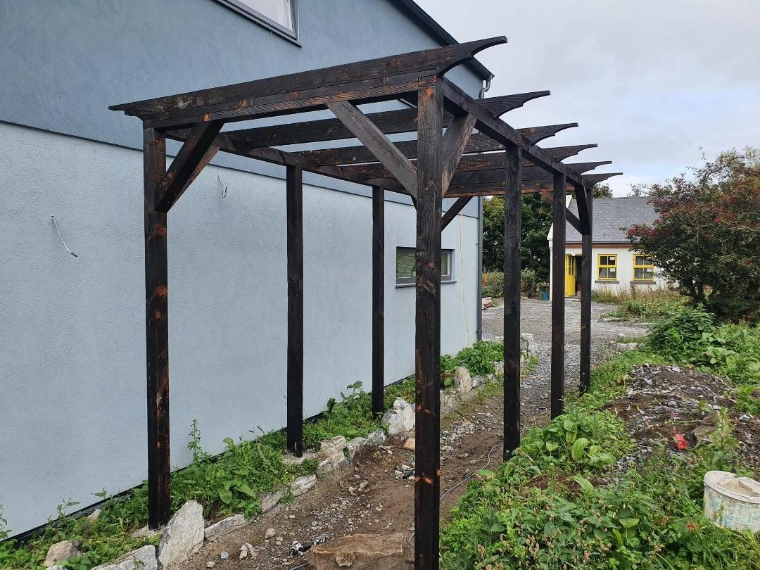 A wooden pergola structure built along the side of a gray house with a few windows, situated in a garden area with rocks, soil, and greenery nearby.