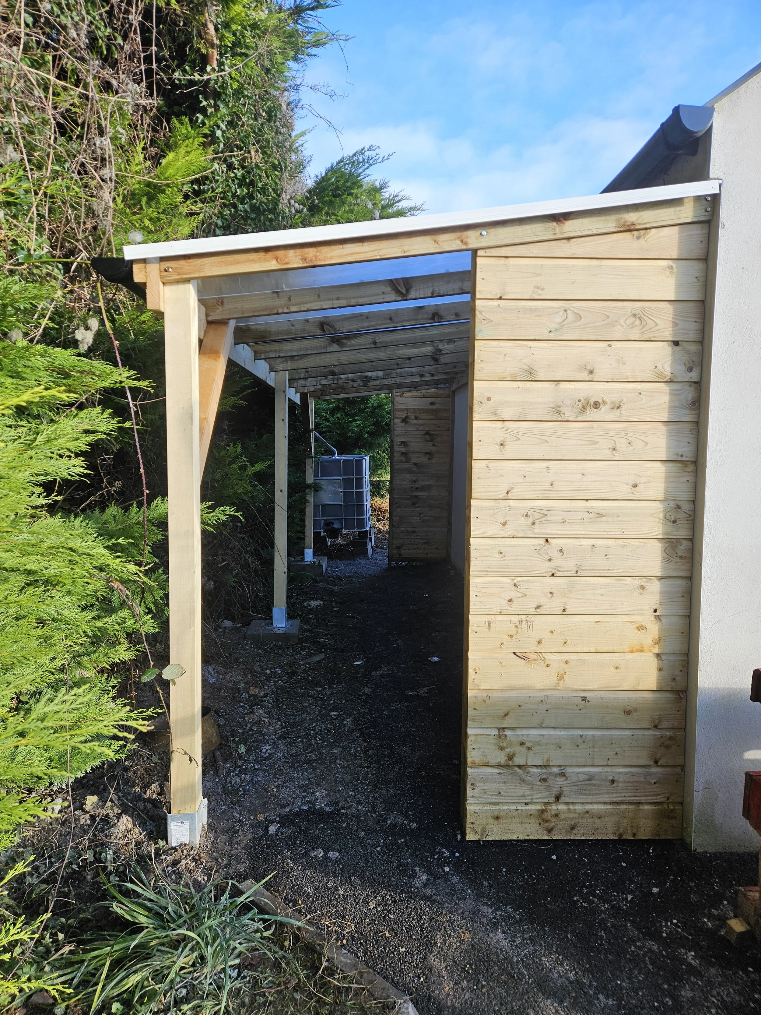 Construction of a wooden structure attached to a white building, with an open side facing a garden and a blue sky with clouds.