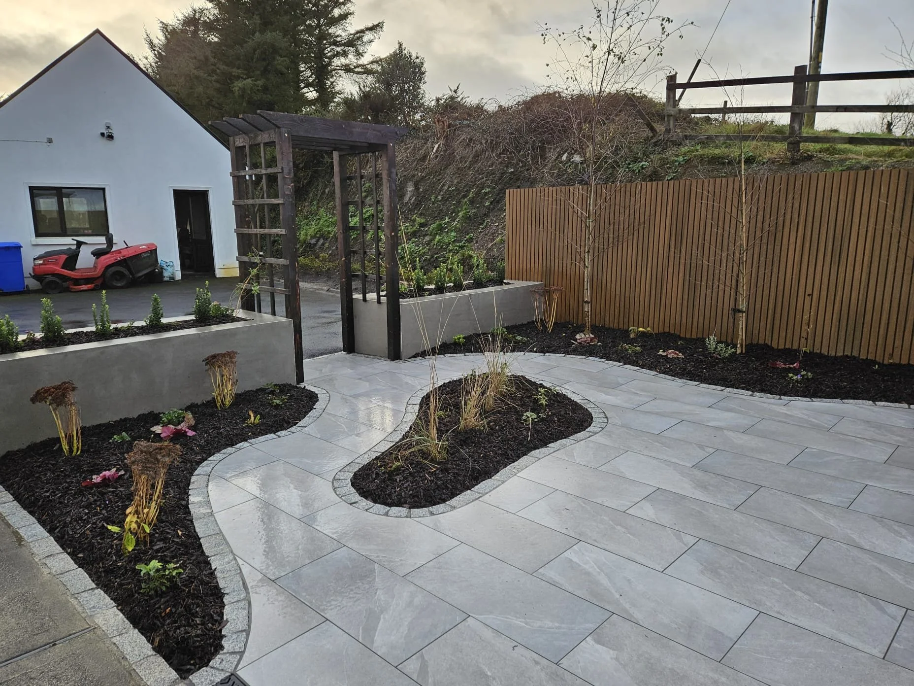 A modern backyard with a paved stone patio, small landscaped garden beds with plants, a wooden garden archway and a wooden privacy fence with trees behind.