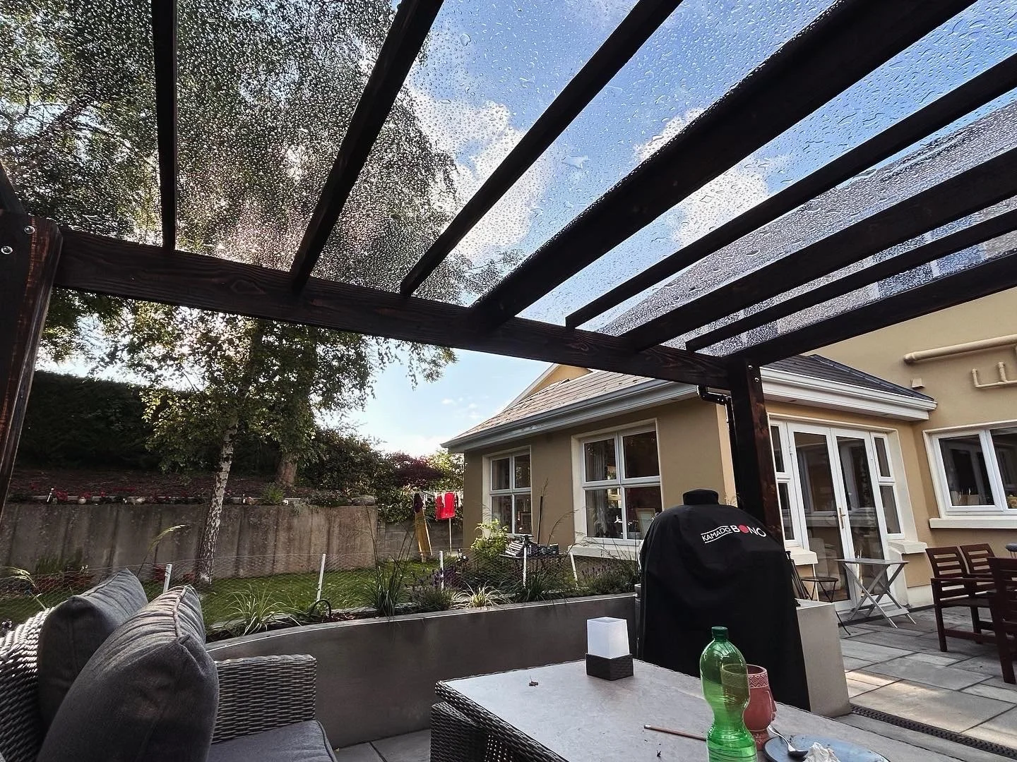 Backyard patio view with a wooden pergola covered in rain droplets, a beige house with large windows, outdoor furniture including a gray cushioned sofa, a white table, and a barbecue grill, garden with trees and a fence, and a clear sky with some clo