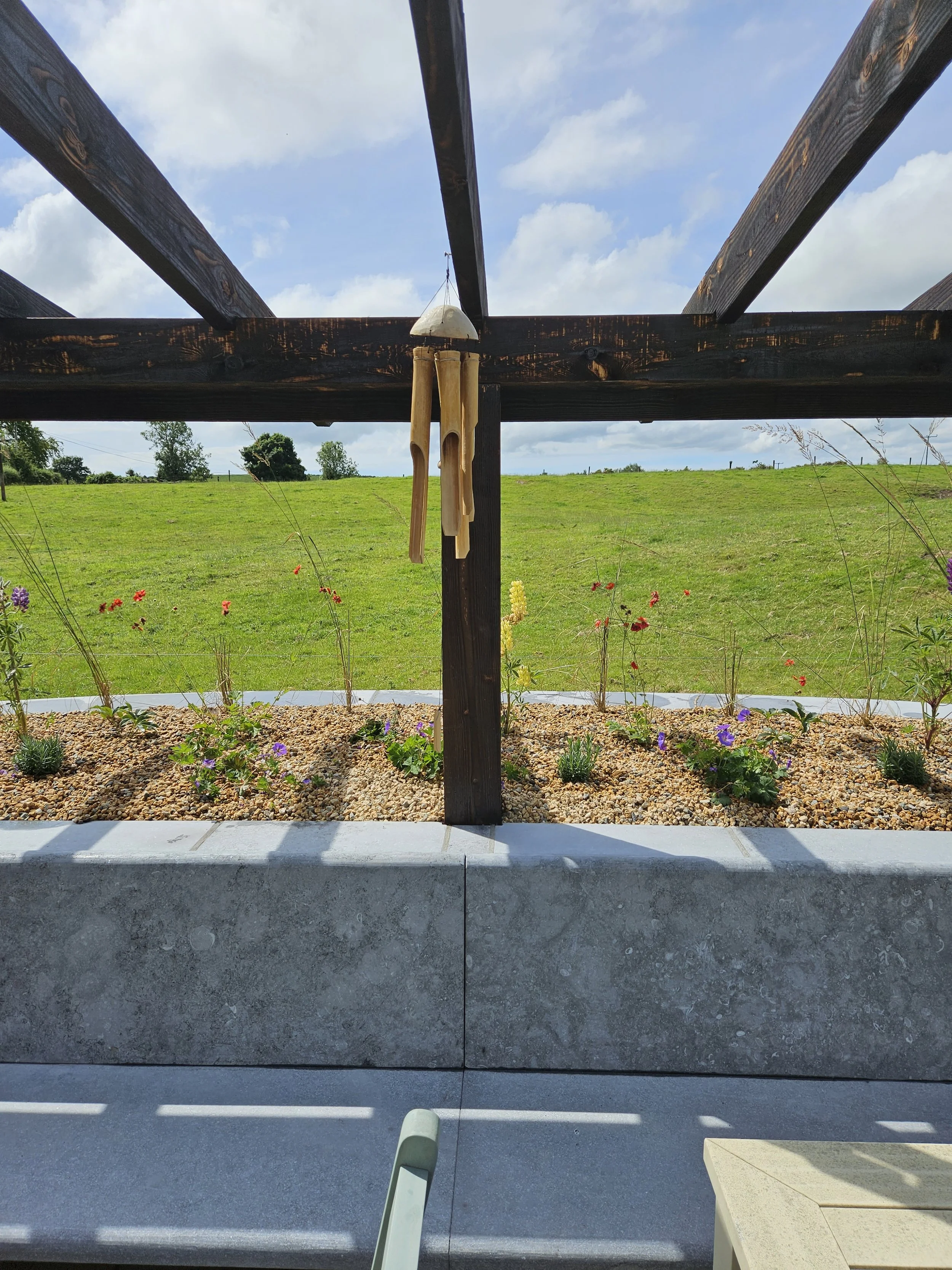 Outdoor garden with a wooden structure, wind chimes hanging, flower bed, green field, blue sky with clouds.