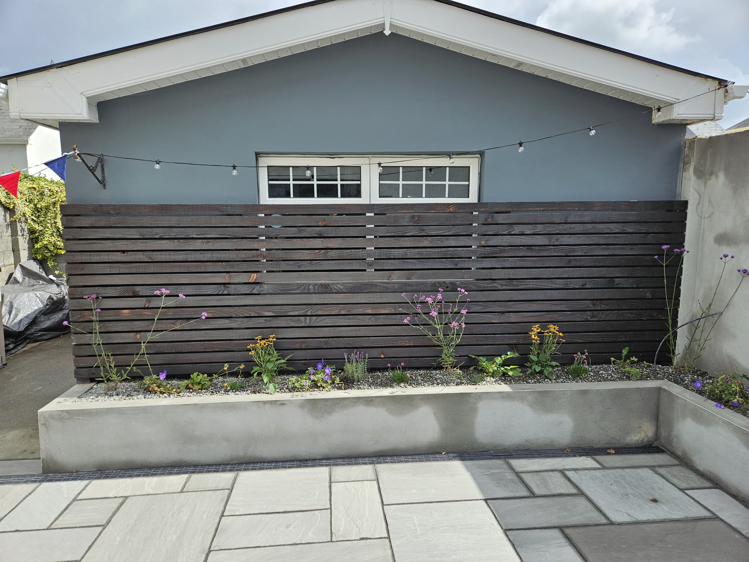 Blue house with white trim, wooden fence, string lights, flower bed with purple and yellow flowers, paved patio, and concrete planter.