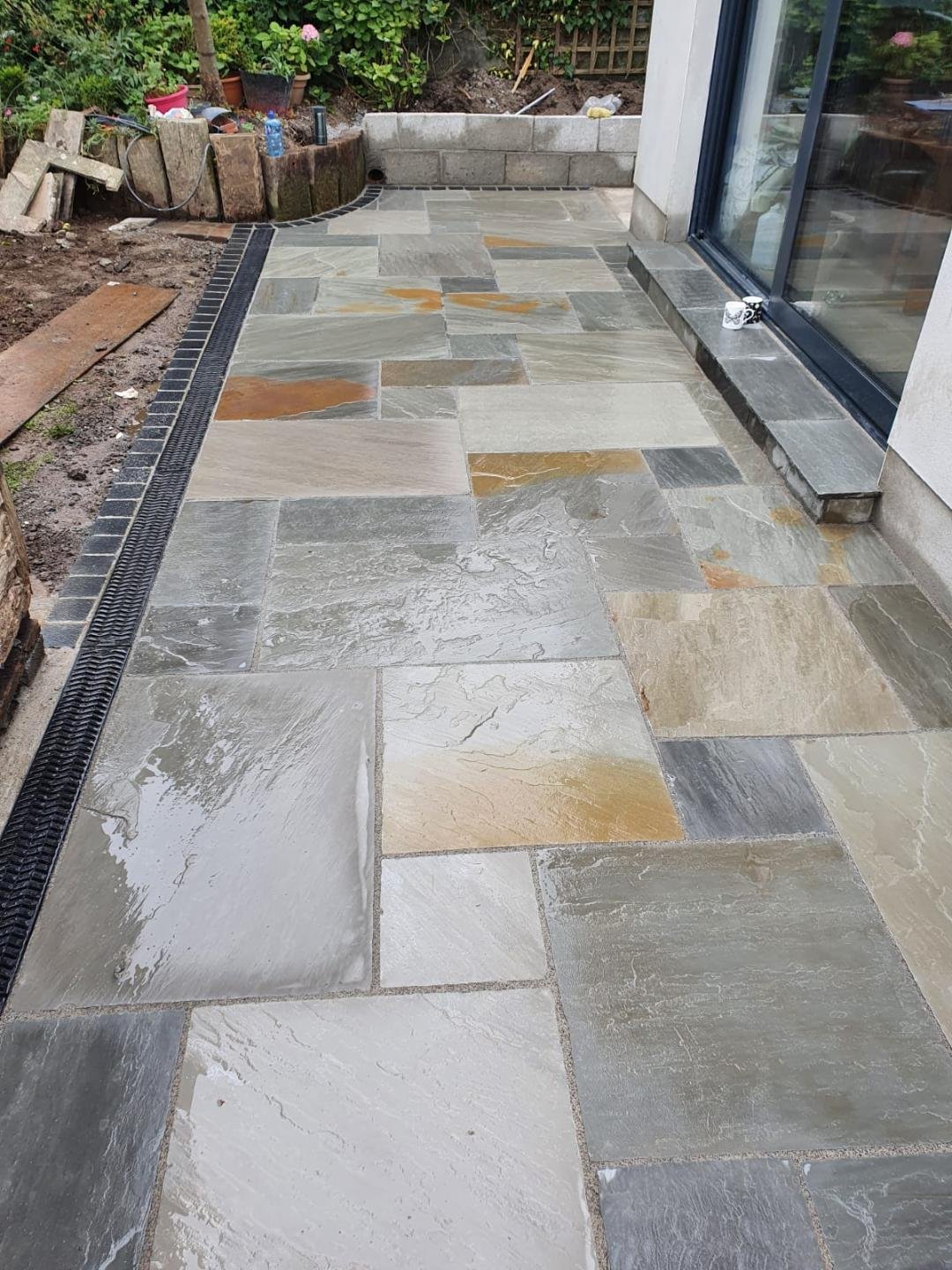 Newly installed concrete patio with large, multi-colored rectangular tiles bordered by small dark tiles, adjacent to a house with large glass sliding doors, and some gardening plants and tools in the background.