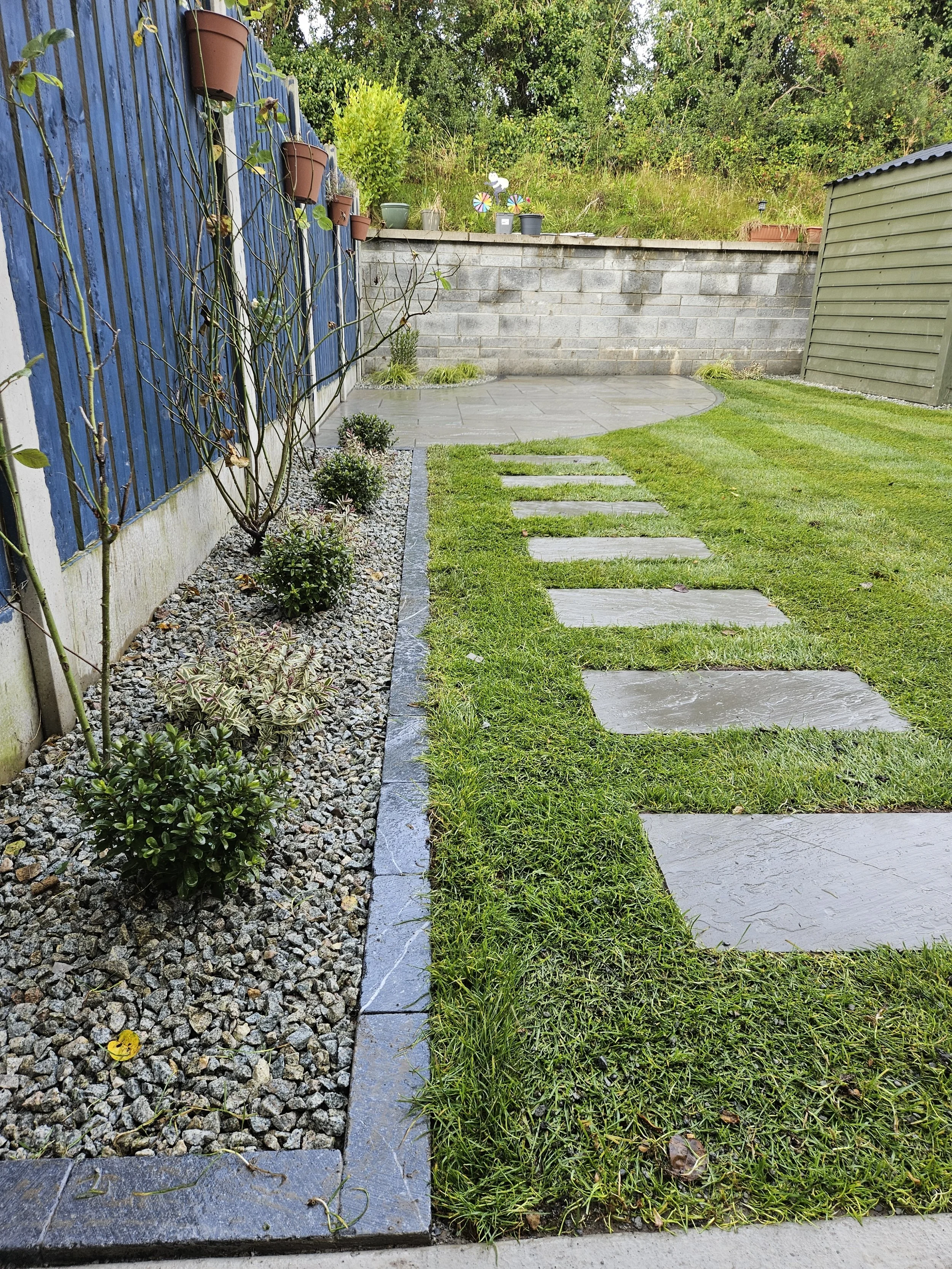 A well-maintained backyard garden with a stone pathway leading to a paved area, bordered by a flower bed with small bushes and gravel, a wooden fence on the left, and a storage shed on the right, with trees and a brick wall at the back.