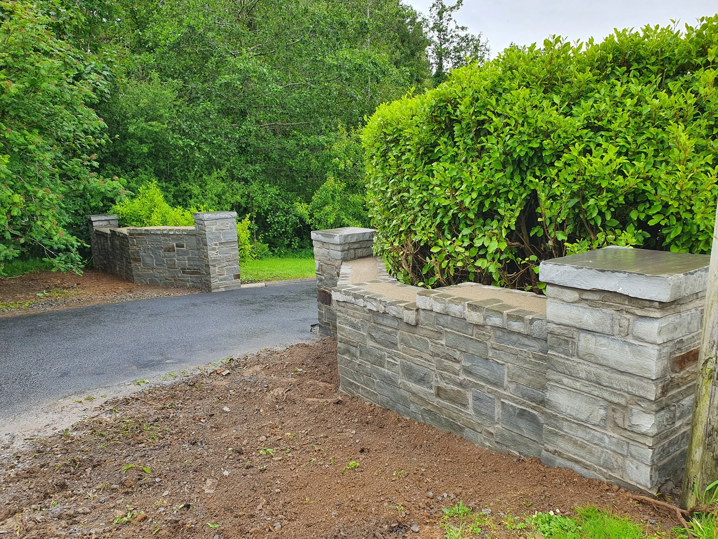 Stone wall and entrance to a paved driveway surrounded by green bushes and trees.