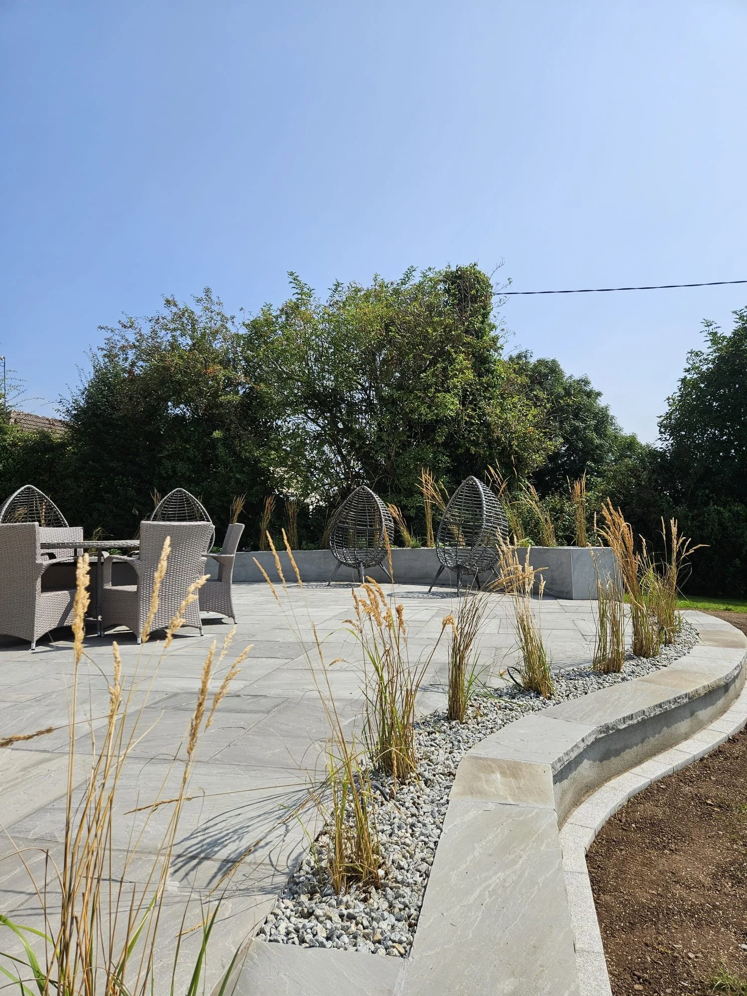 A modern outdoor patio with woven chairs, a round table, and decorative grass plants along a curved concrete border, with lush green trees and a clear blue sky in the background.