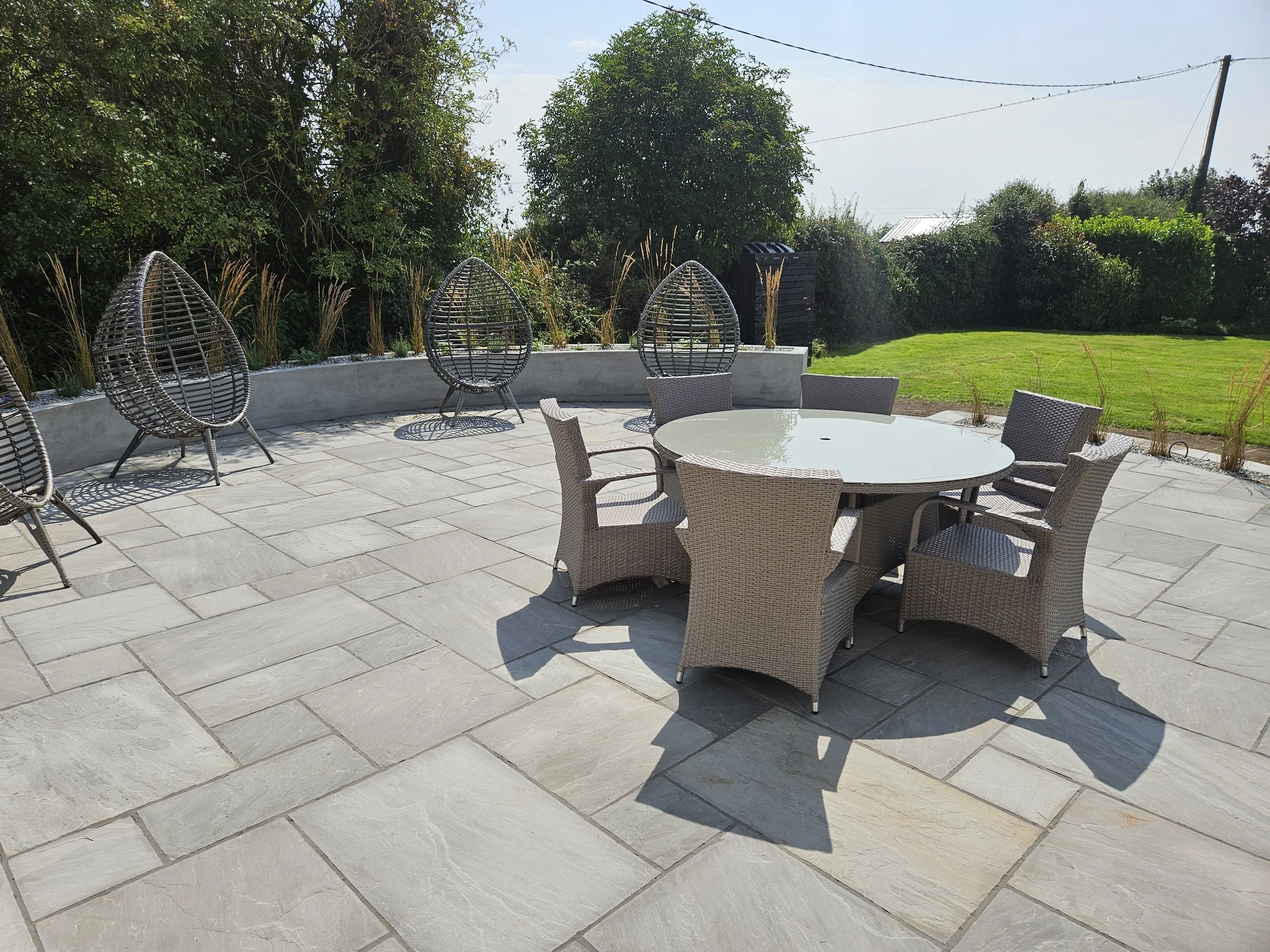 A backyard patio with a round glass table surrounded by six wicker chairs, and four teardrop-shaped wicker lounge chairs in front of a curved concrete planter with plants, green lawn, trees, and bushes in the background.