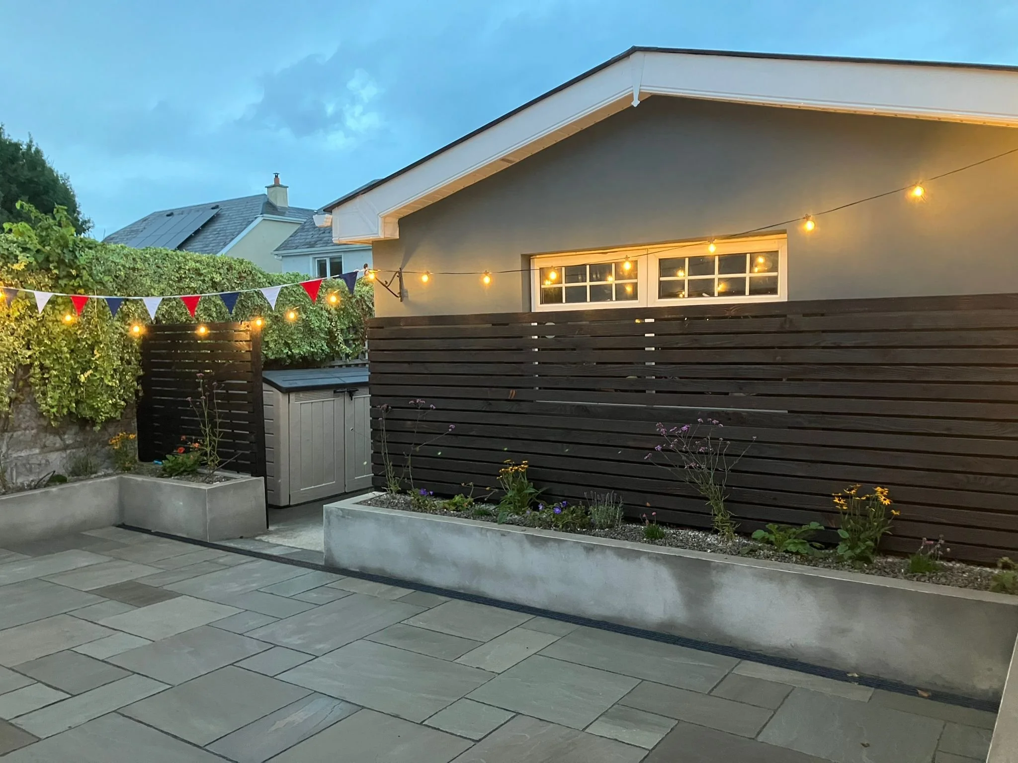 A backyard patio scene at dusk with string lights and patriotic bunting. The patio has stone paving, a garden with flowers, and a dark wooden fence. The background shows neighboring houses and a cloudy sky.