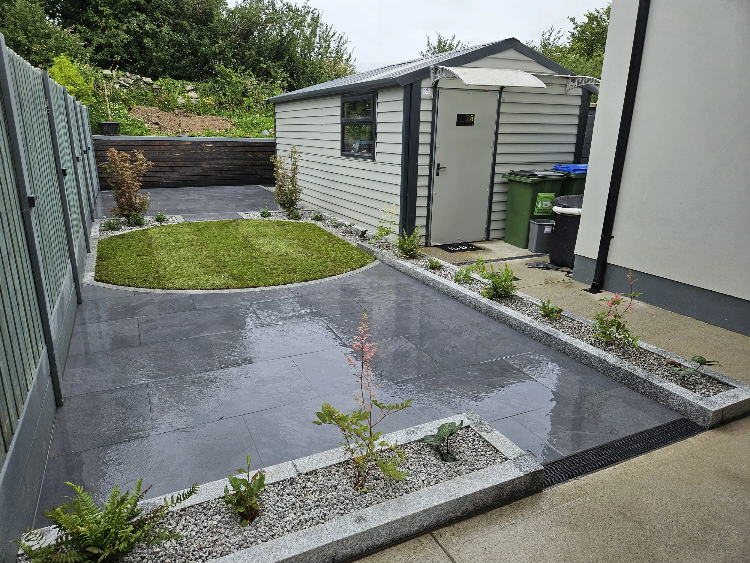 Rain-soaked patio with gray tiles, a small grass patch, and a row of plants along the edge near a shed with trash bins, surrounded by green trees and a fenced area.