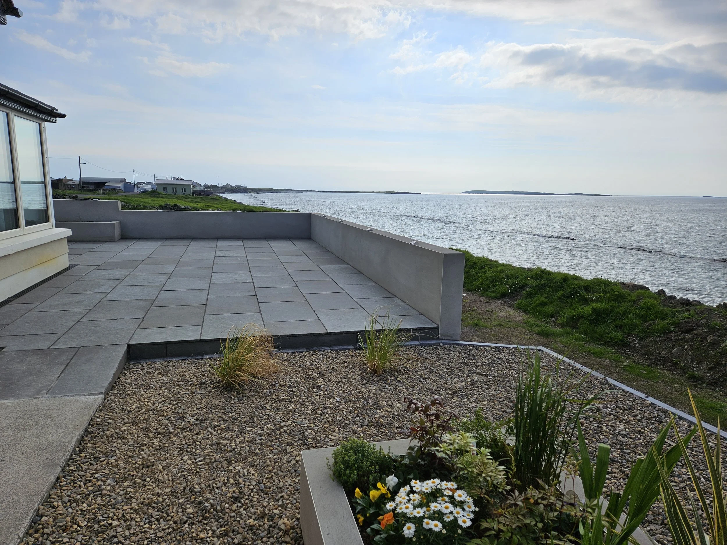 View of a seaside patio with a concrete tiled floor, a low wall, and a garden bed with flowers and plants, overlooking the ocean with a partly cloudy sky.