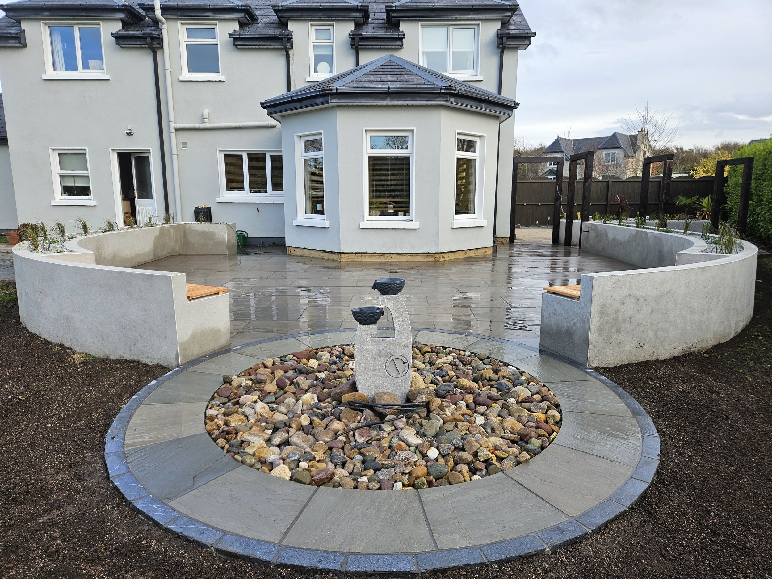 Modern backyard with curved concrete benches, a central stone fountain, and a patio area in front of a house with multiple windows and a small turret, wet from recent rain.