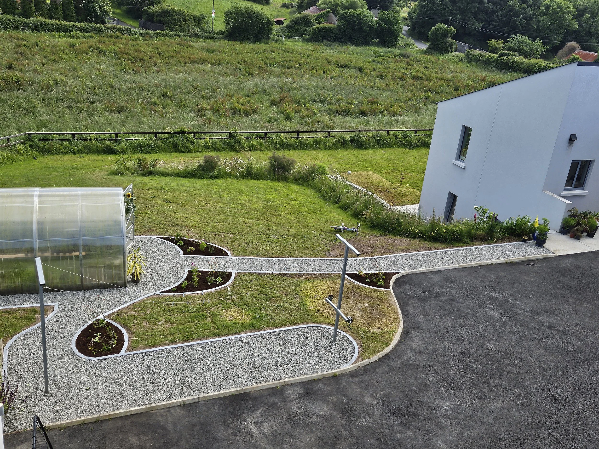 A modern white house with a garden and a greenhouse, gravel pathways, sunflowers, and potted plants, in a rural setting with green fields and trees.
