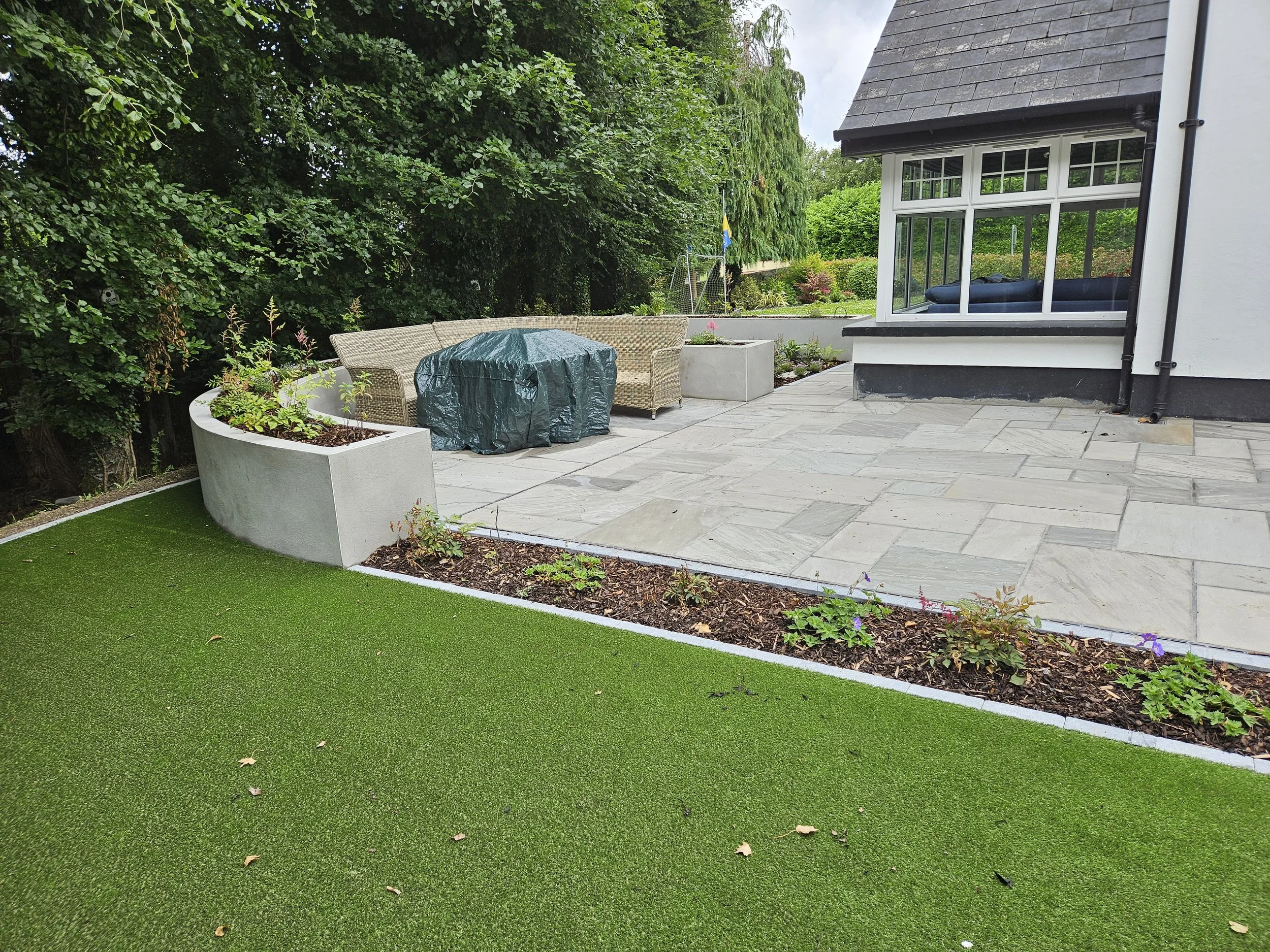 Backyard patio with a raised flower bed, outdoor seating, and a covered round table, adjacent to a house with a bay window and garden area.