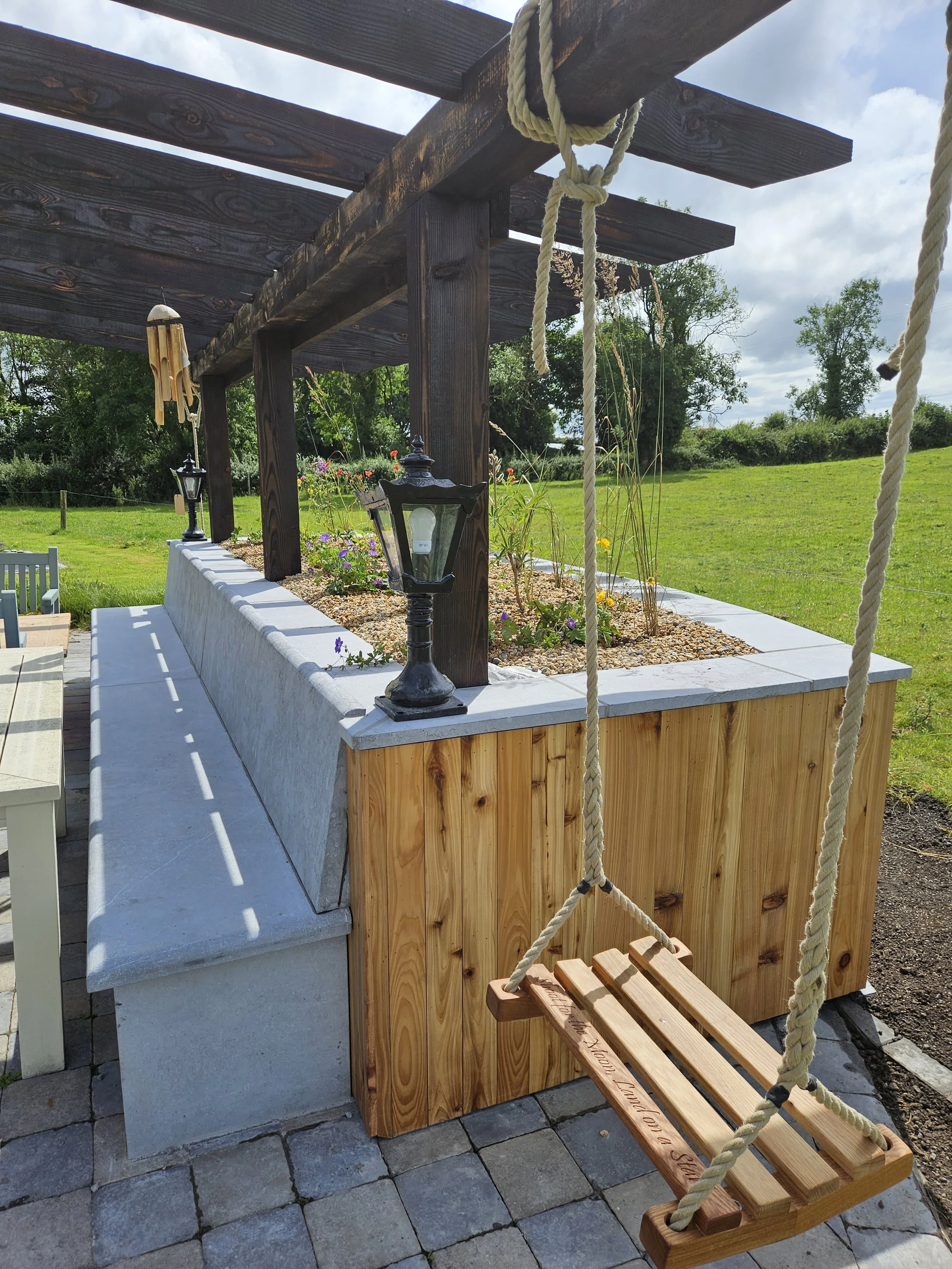 A raised garden bed with flowers, a wooden pergola overhead, and a wooden swing with rope hanging from the pergola in a backyard.