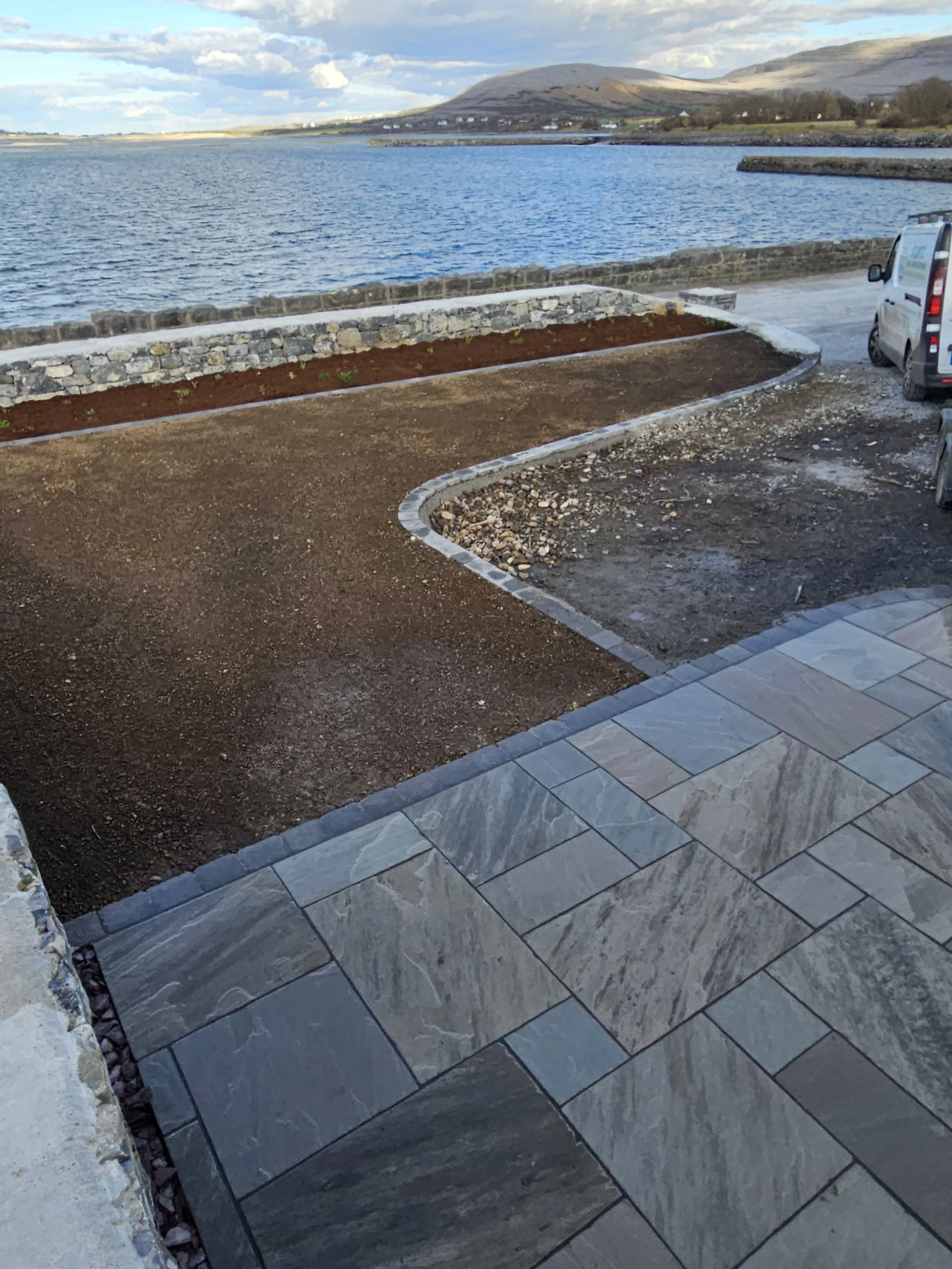 Partly finished patio with large stone tiles, a gravel area, and a flower bed with a stone border by a body of water, with hills in the background and parked vans to the right.