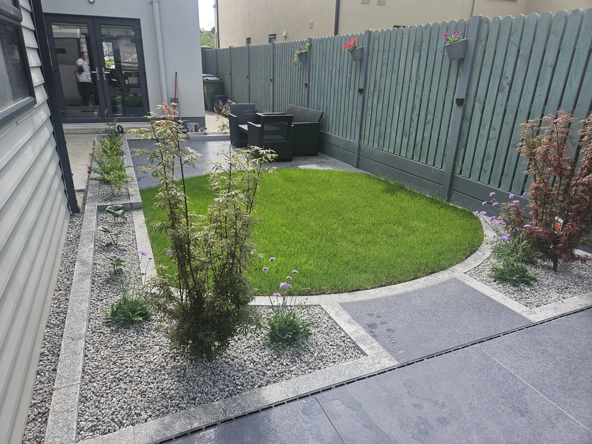 A backyard with a small lawn, landscaped with gravel borders, plants, and flowers. There is a paved area with a sitting arrangement of black outdoor furniture near a green wooden fence with flower baskets.