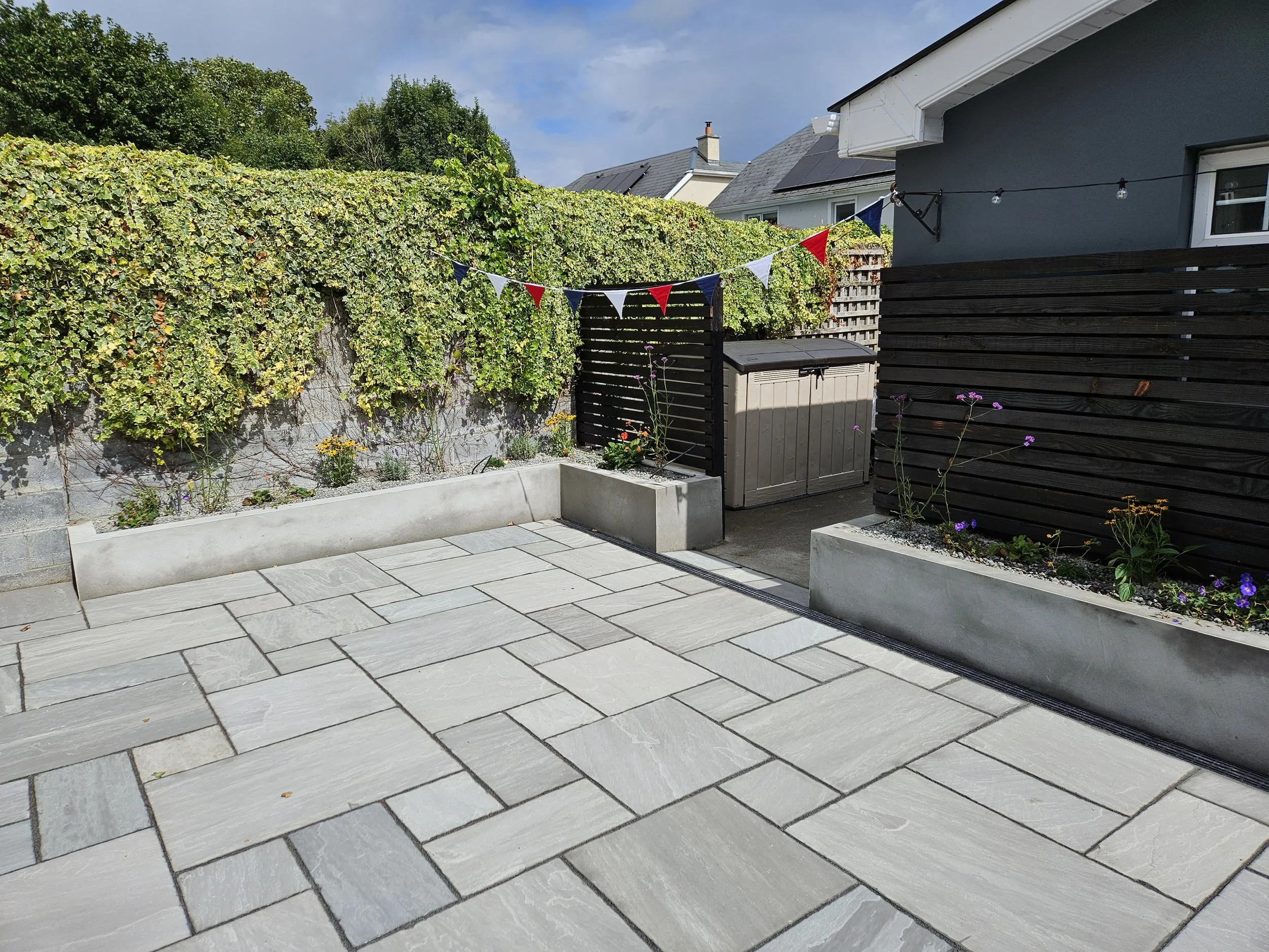 A backyard patio with light-colored stone paving, bordered by raised flower beds with various plants and flowers. A string of festive triangular banners in red, white, and blue hangs across the patio, and there is a gray storage shed next to a dark b