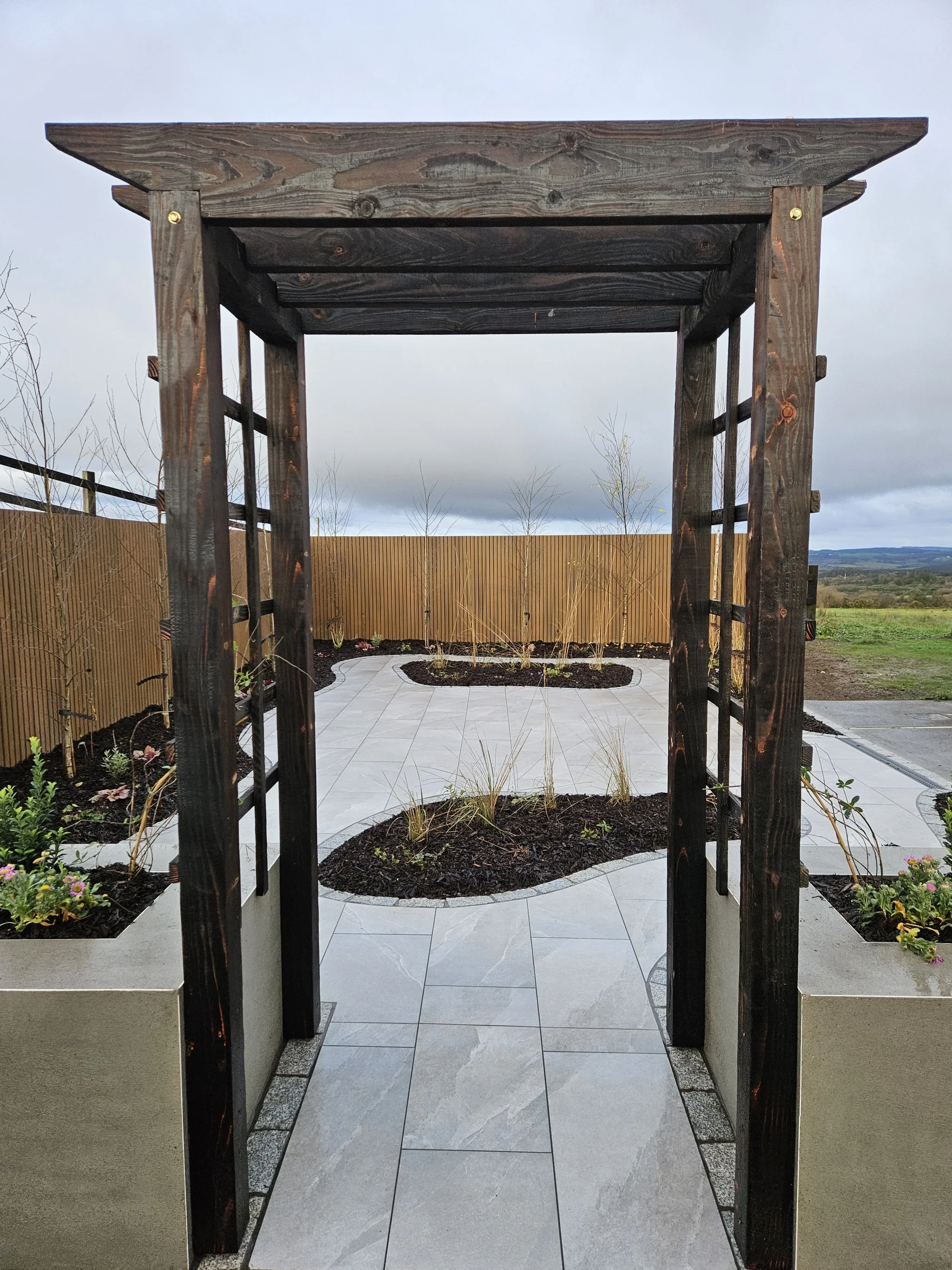 Wooden garden arbor over a stone-paved pathway leading to a landscaped backyard with small trees and a wooden fence, under an overcast sky.