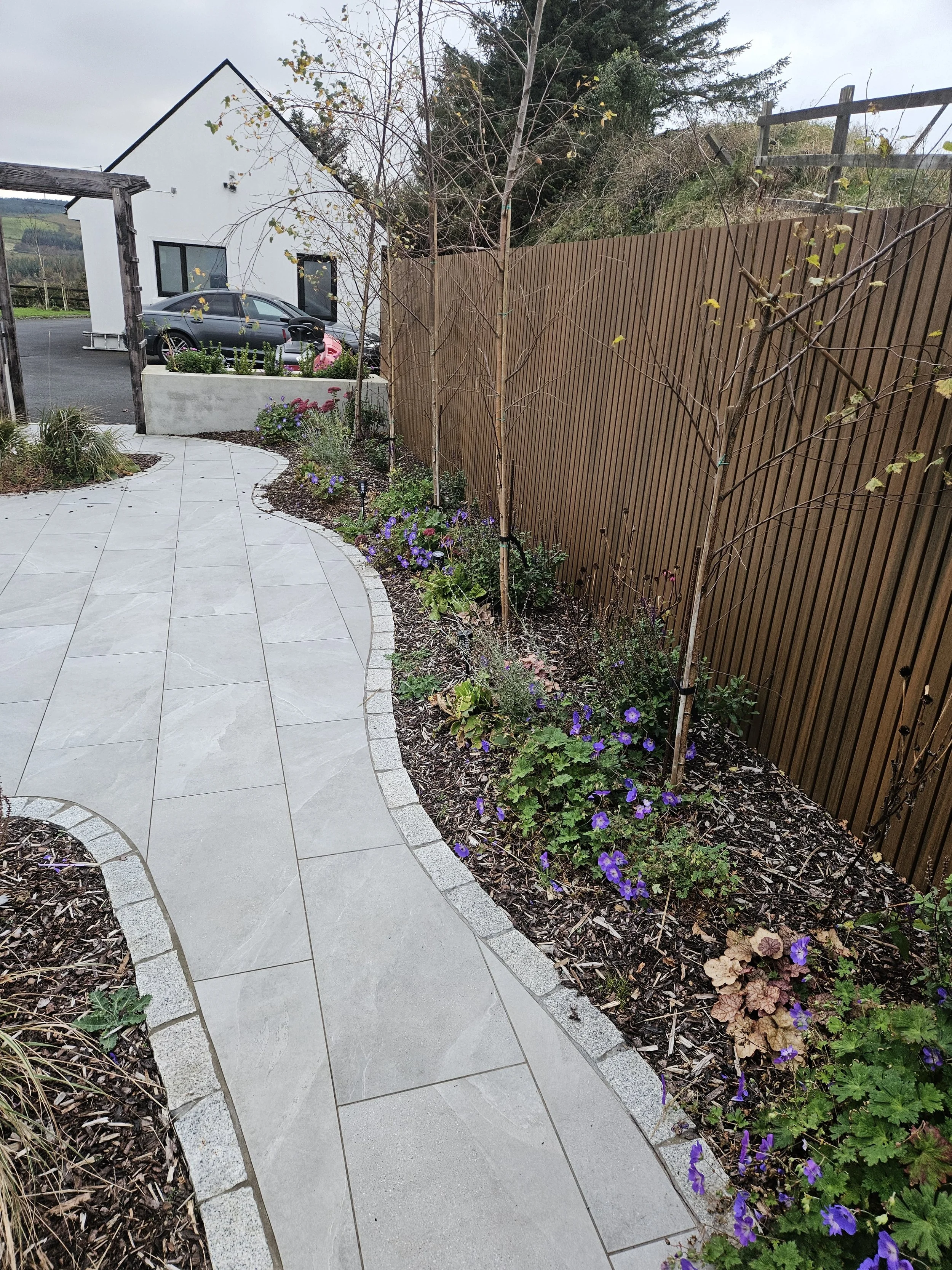 A curved stone pathway bordered by a garden with purple flowers and small trees, with a wooden fence on one side and a parking lot and white building in the background.