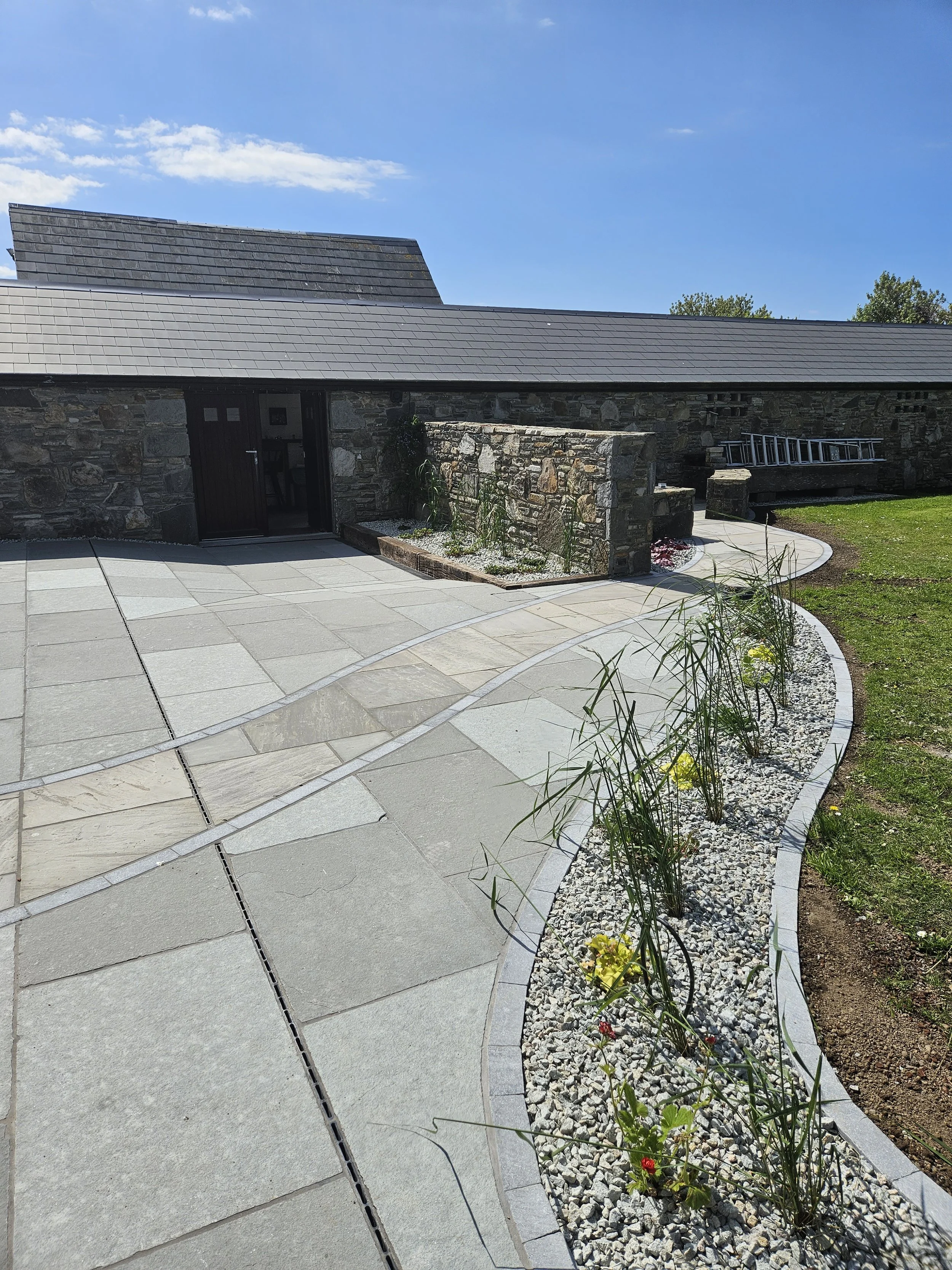 A modern garden pathway made of large stone tiles with a curved shape, bordered by gravel and small plants. There is a stone building with a slate roof in the background and a ladder resting against the building. The sky is clear and blue.