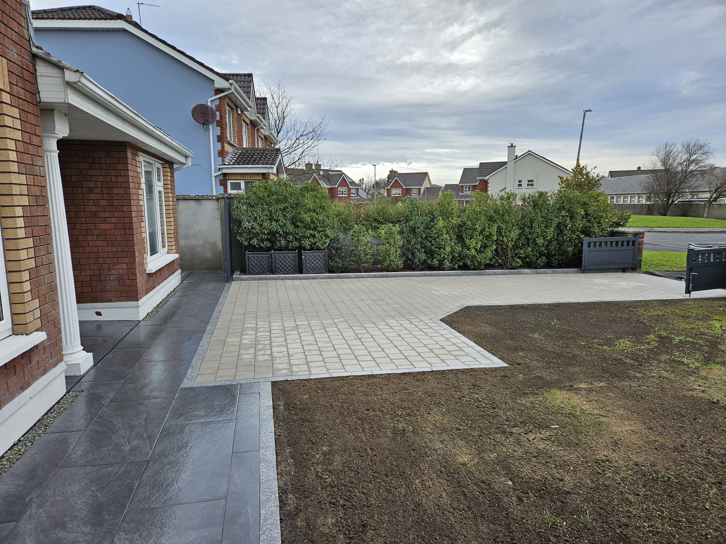 View of a constructed outdoor patio area next to a brick house with a window, featuring gray stone tiles and a brick pavement area, with a garden bed and bushes in the background under a cloudy sky.