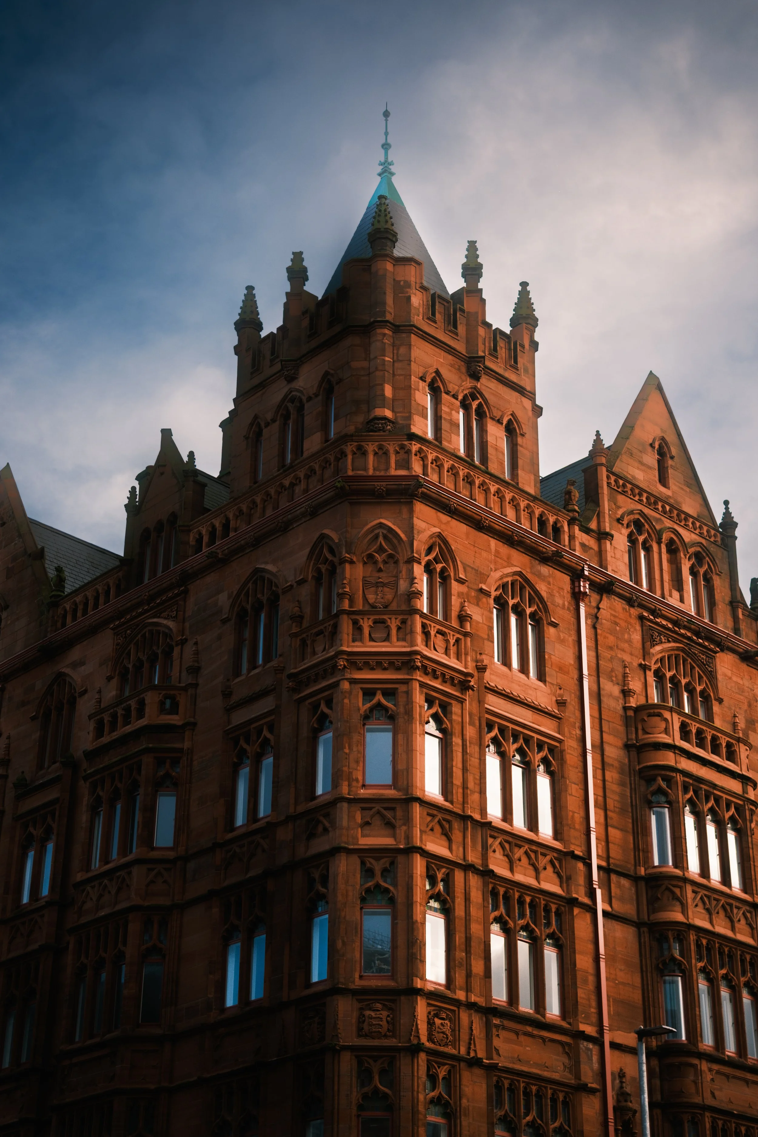 A historic, castle-like building made of brown sandstone with ornate architectural details, pointed turrets, and numerous arched windows, under a partially cloudy sky.