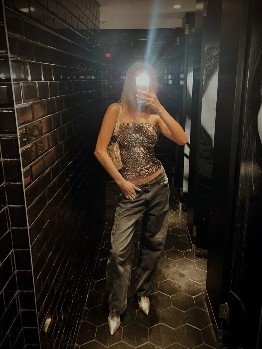A woman taking a mirror selfie in a bathroom with black tiled walls and hexagonal floor tiles. She is wearing a sparkly strapless top, loose gray jeans, and pointed shoes. Her face is obscured by her phone flash.