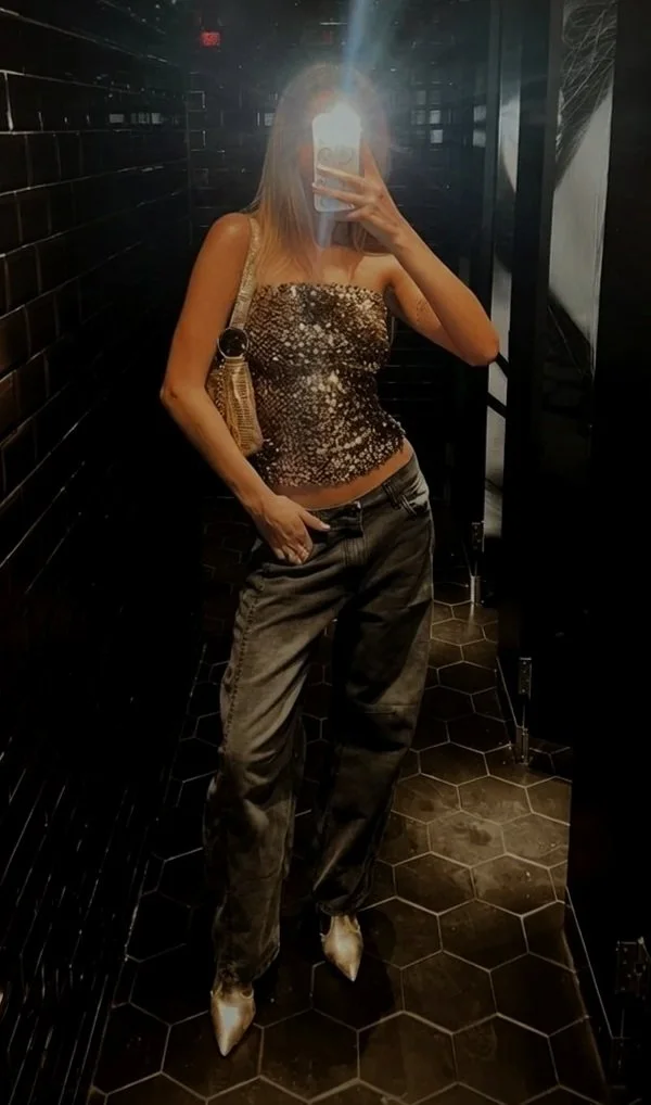 Woman taking a mirror selfie in a dimly lit room with black tile walls and hexagon floor tiles. She is wearing a sleeveless, sparkly top, baggy pants, and pointed shoes. She has a shoulder bag.