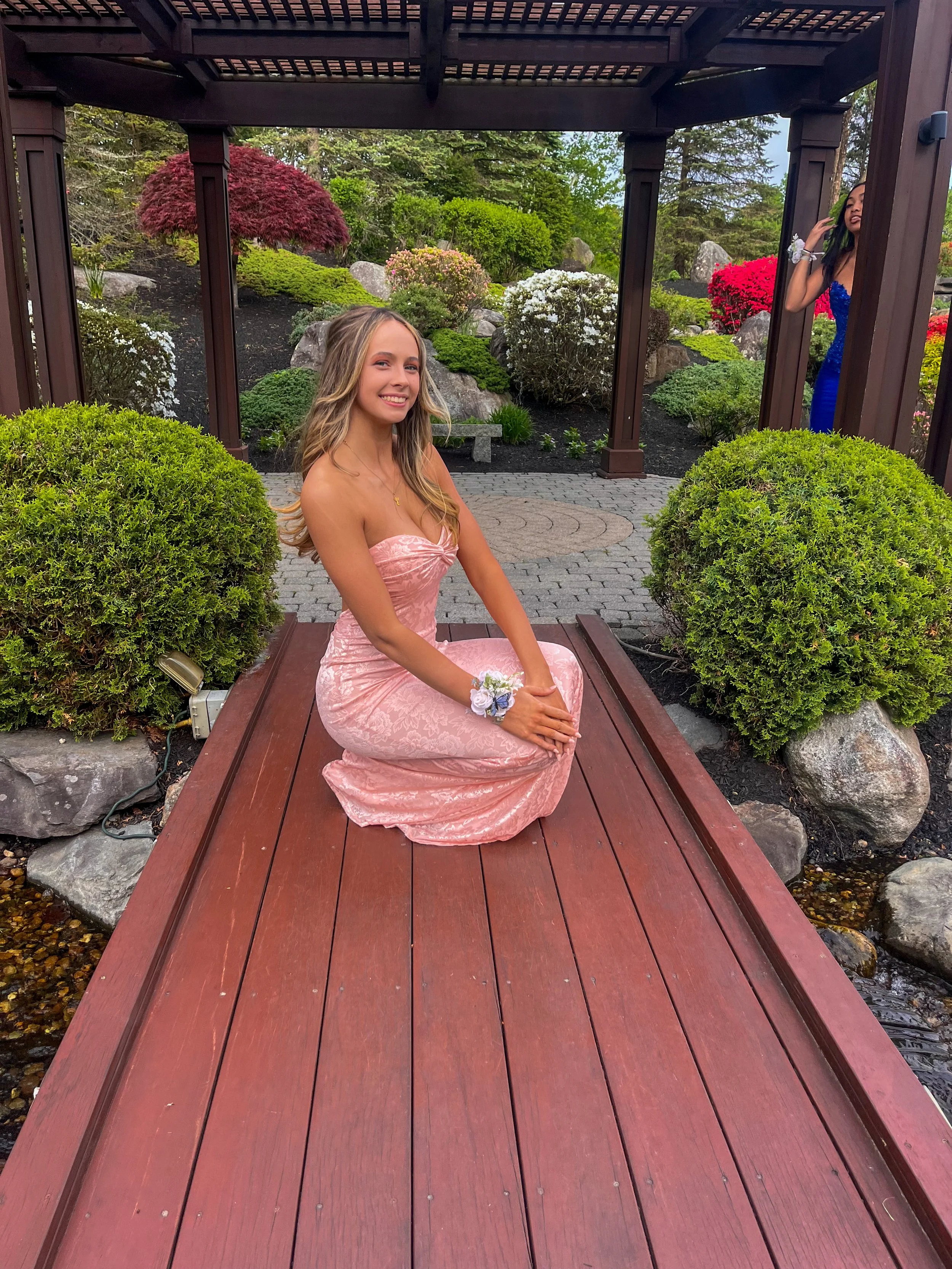 A young woman in a pink strapless dress sitting on a wooden bridge in a garden with colorful plants and bushes, while another woman in a blue dress stands behind a wooden structure in the background.