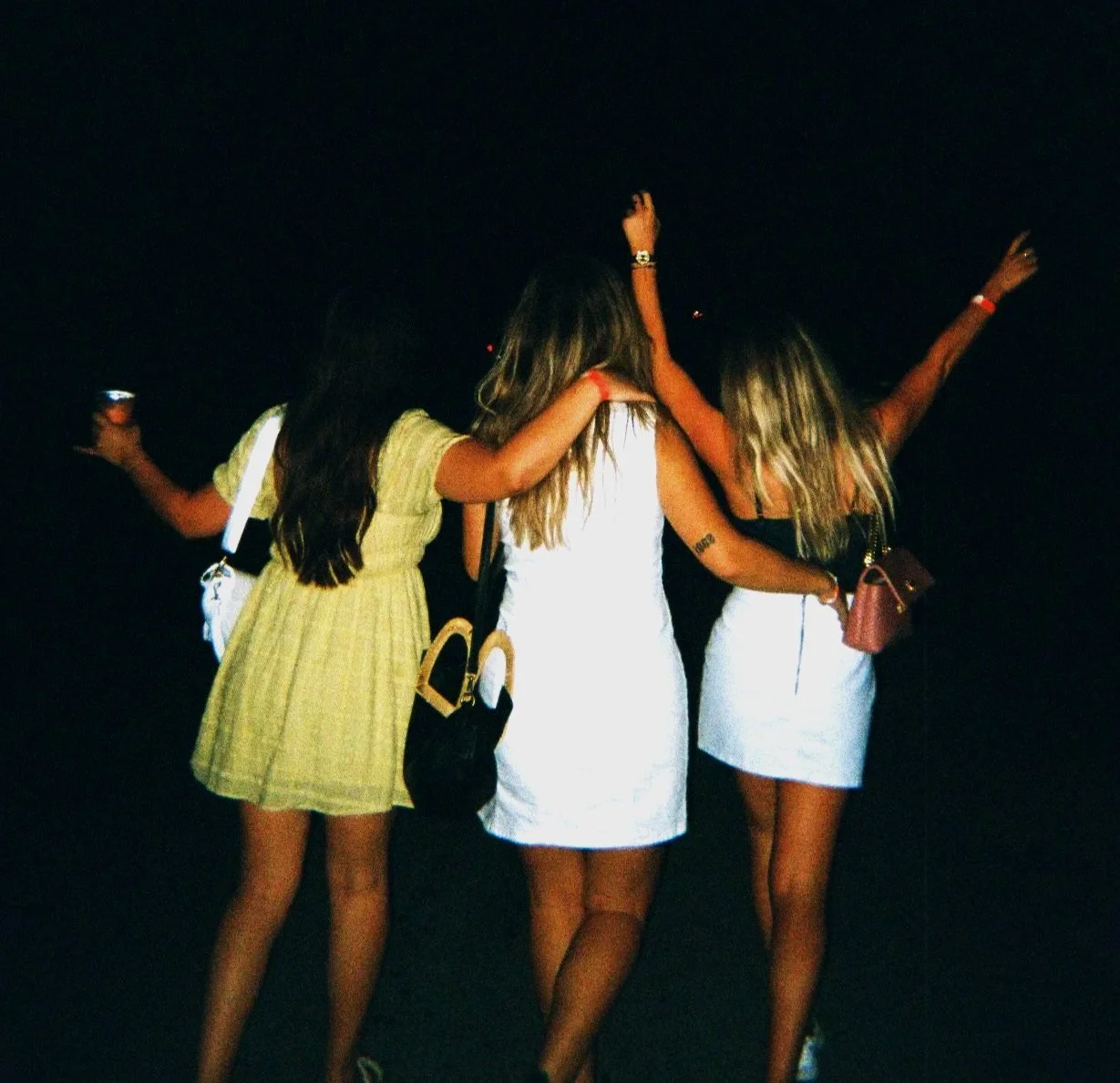 Three women with long hair wearing summer dresses at night, standing close together with arms around each other, facing away from the camera.