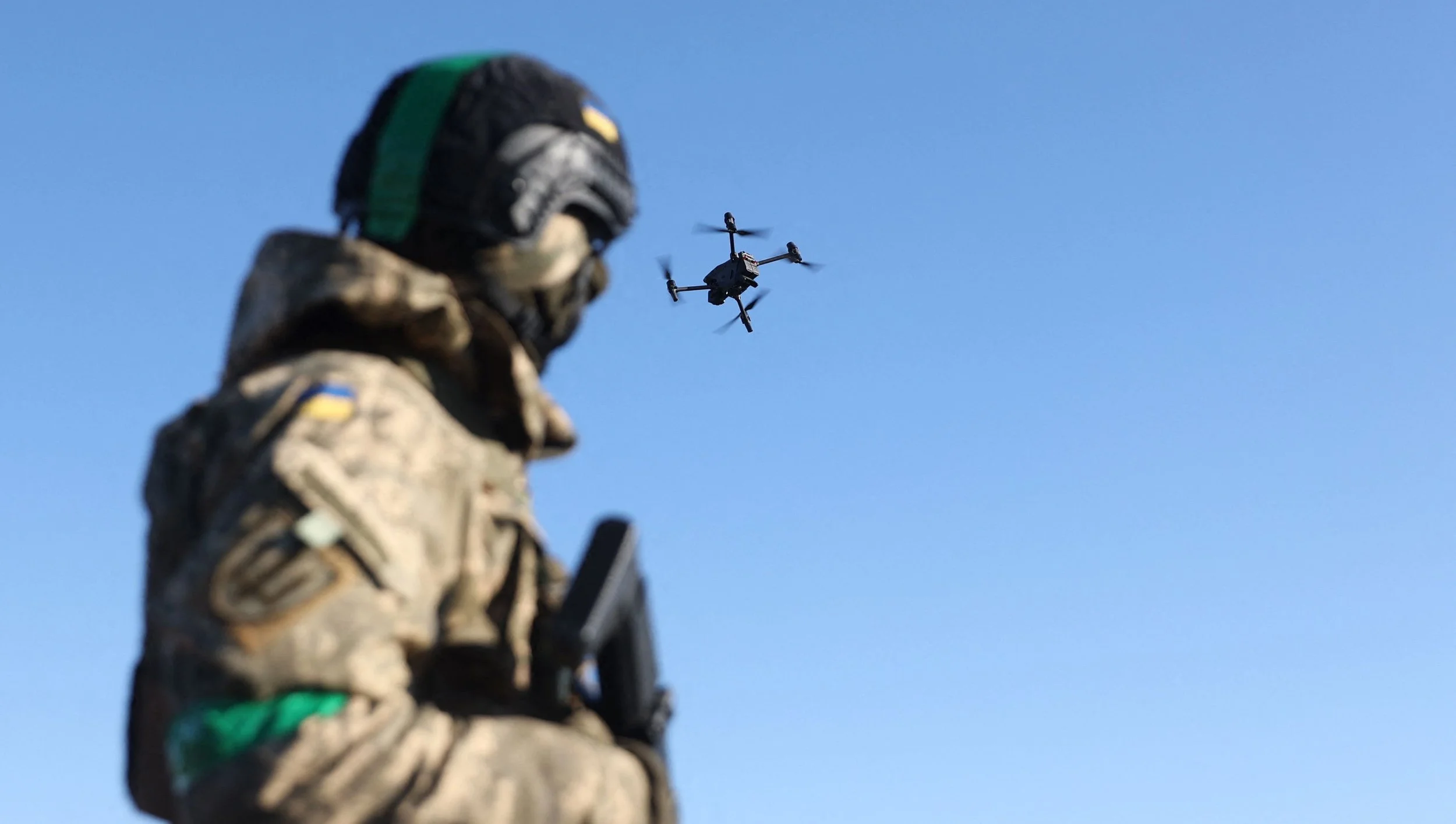 A soldier in camouflage uniform and helmet holding a weapon, standing against a clear blue sky with a drone flying overhead.