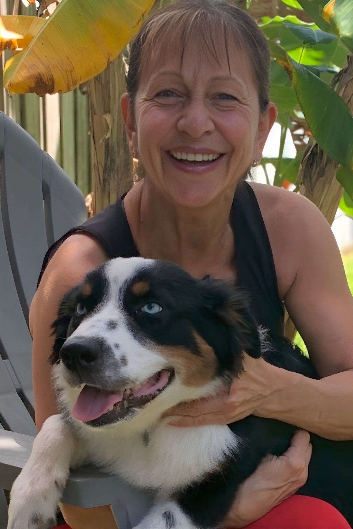 A smiling woman holding a black and white Australian Shepherd puppy with blue eyes outdoors among green plants.