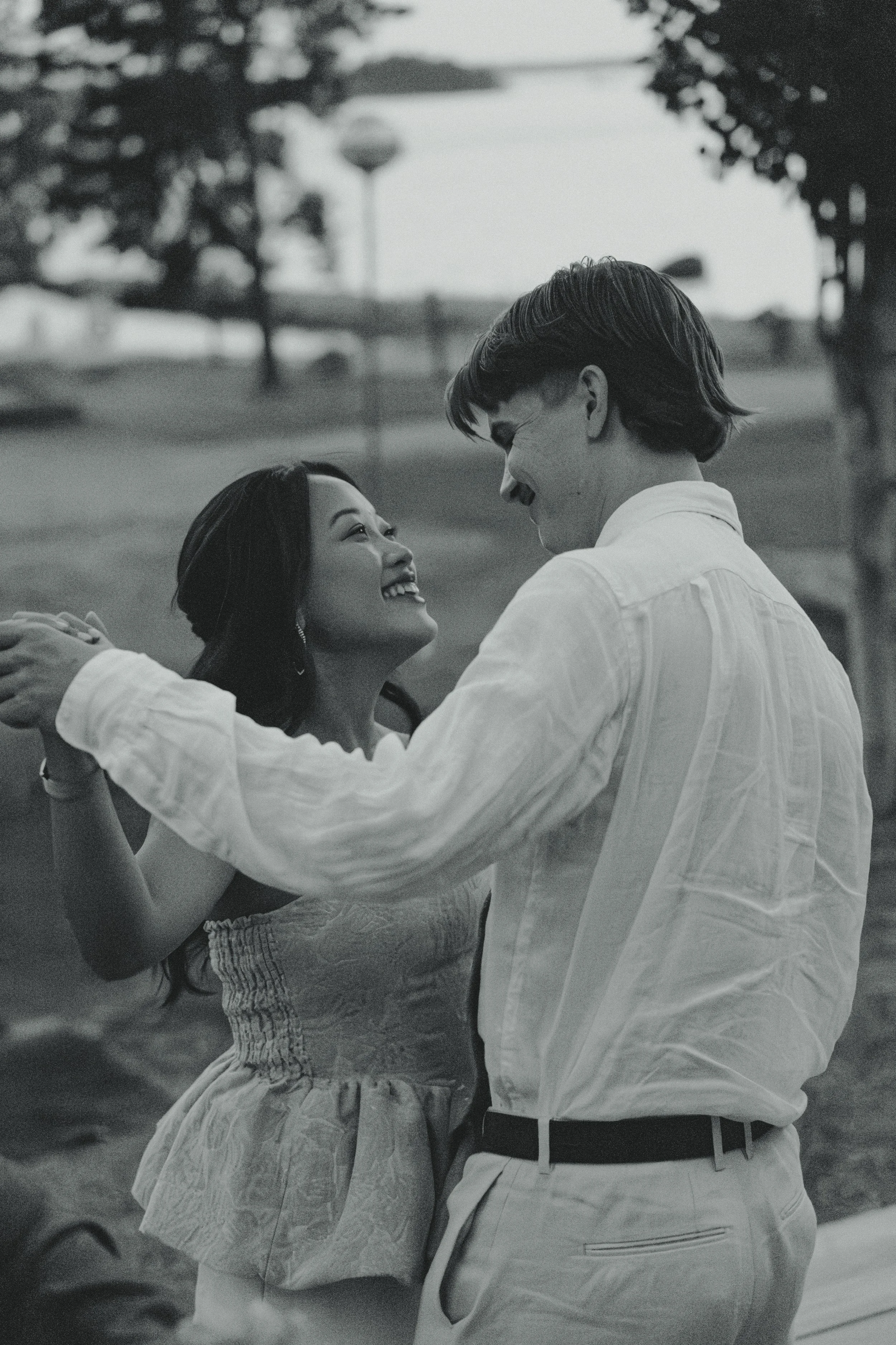 A black-and-white photo of a couple dancing outdoors, smiling and looking into each other's eyes, with a park and trees in the background.