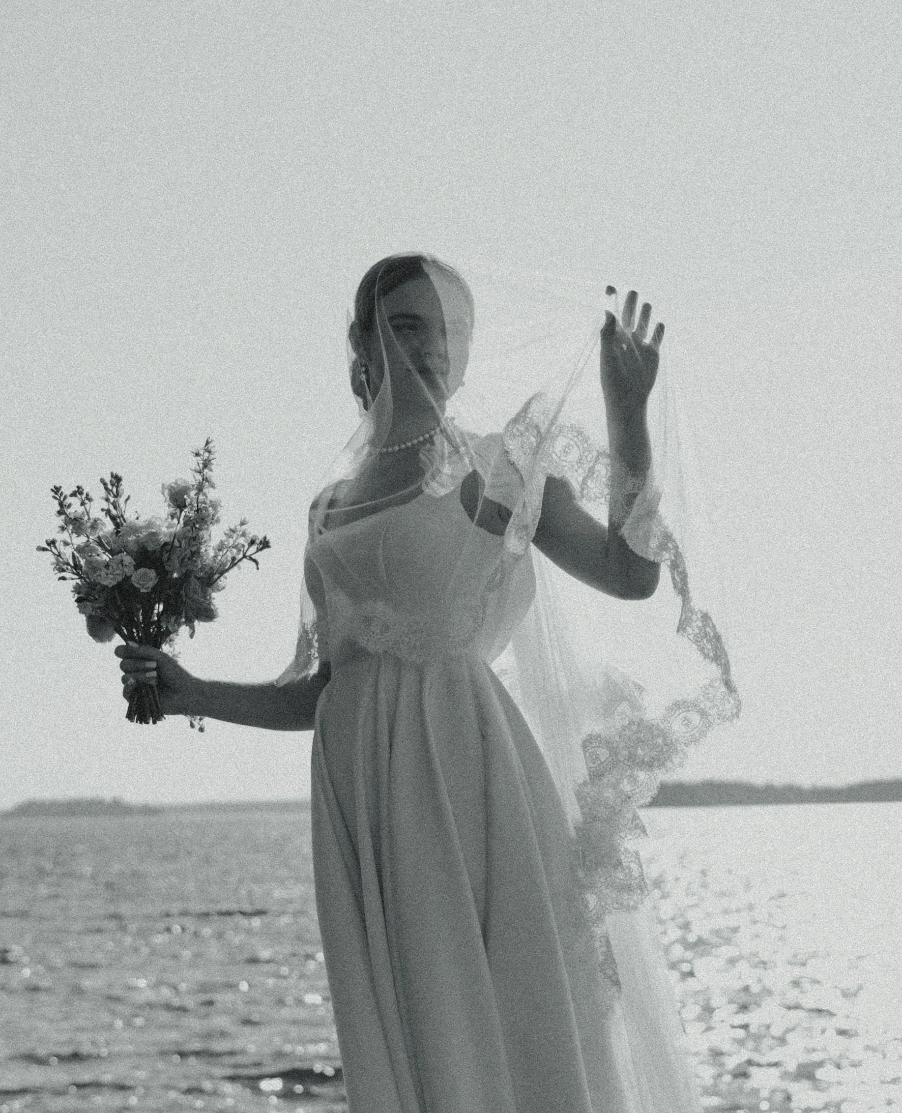 A woman in a wedding dress holding a bouquet of flowers, standing outdoors behind a sheer veil, with water and a distant shoreline in the background.