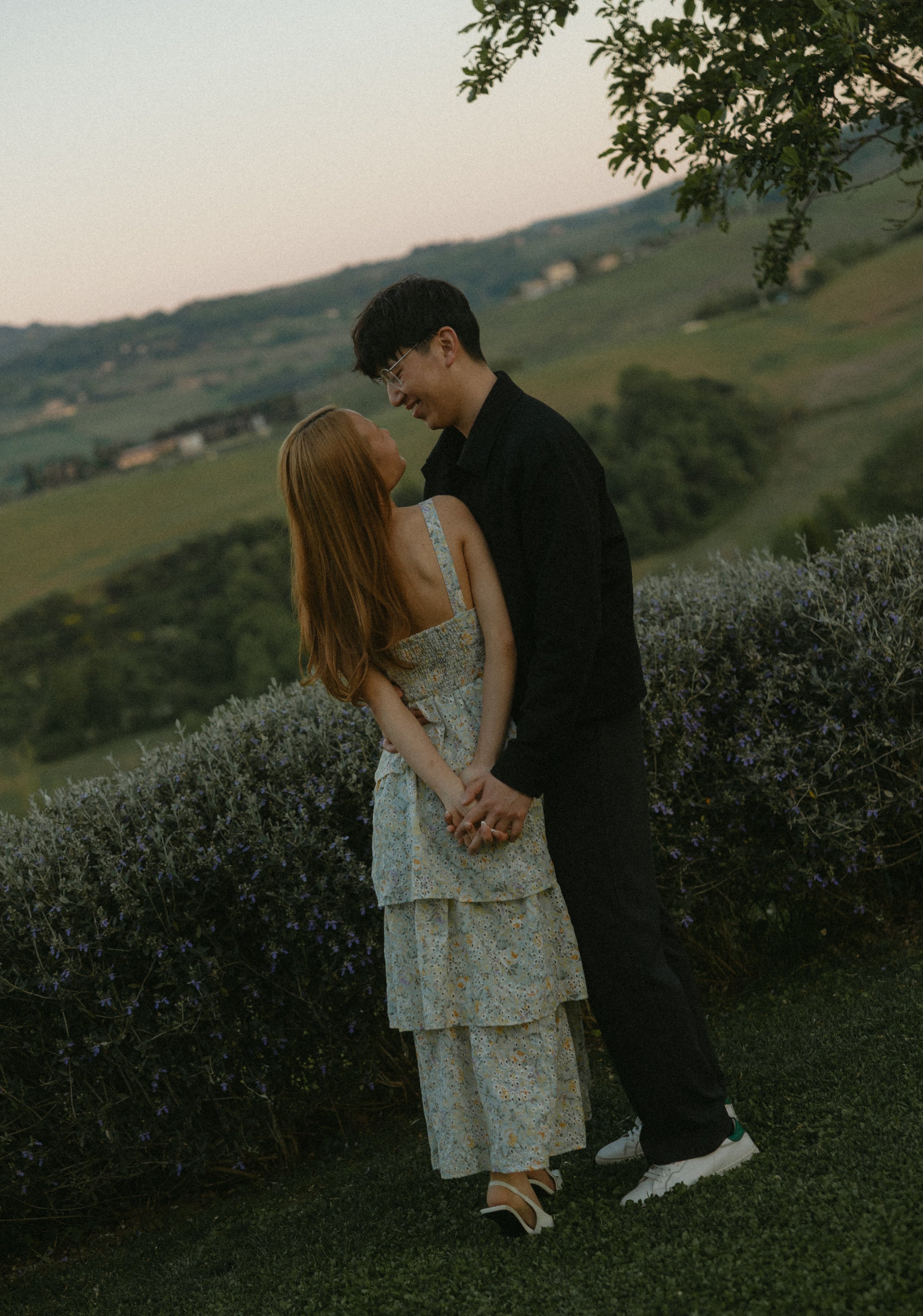 A couple standing close, holding hands, outdoors in a scenic landscape with rolling hills and a tree, during sunset.