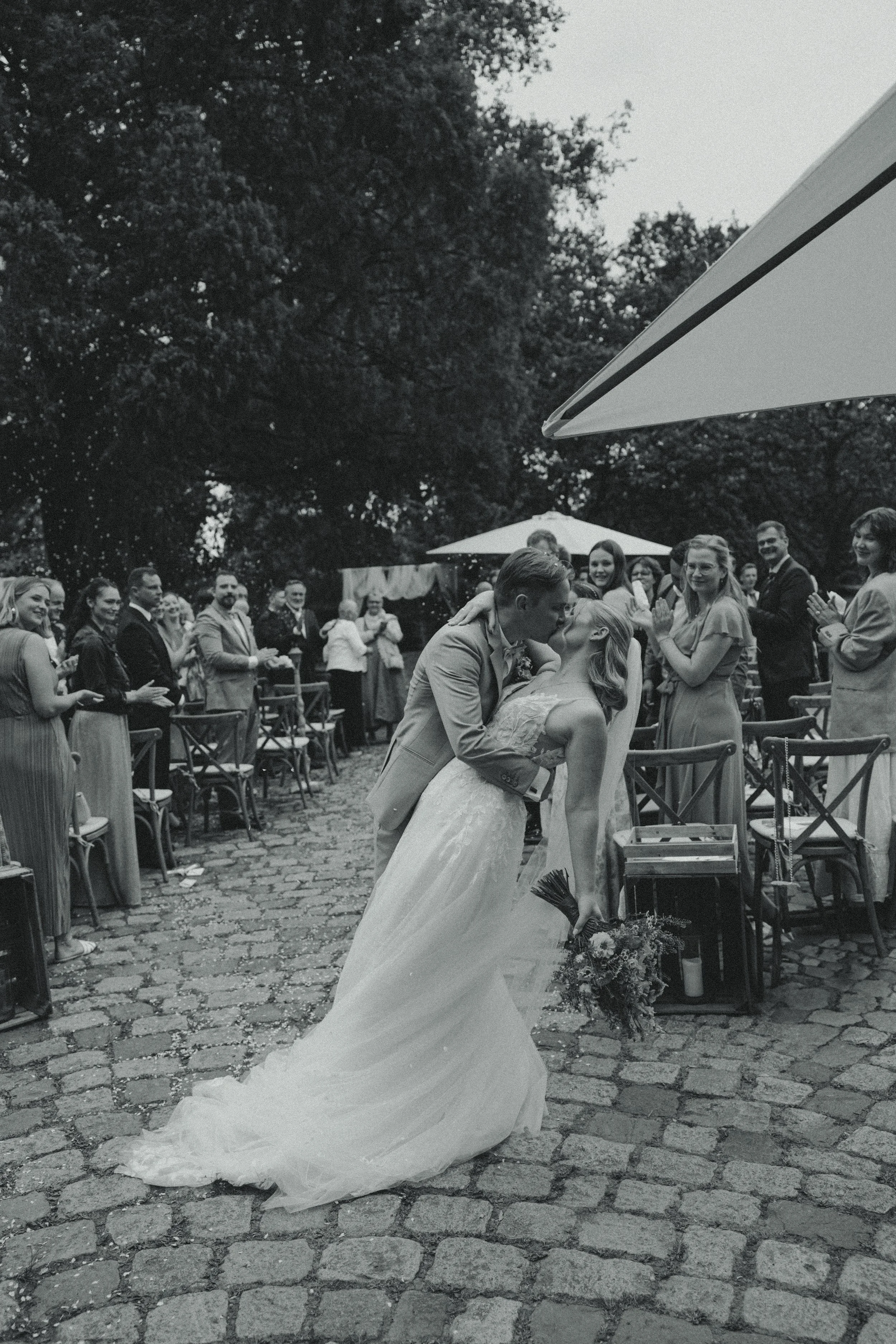 A black-and-white photo of a newlywed couple kissing at an outdoor wedding reception with guests clapping and smiling around them.