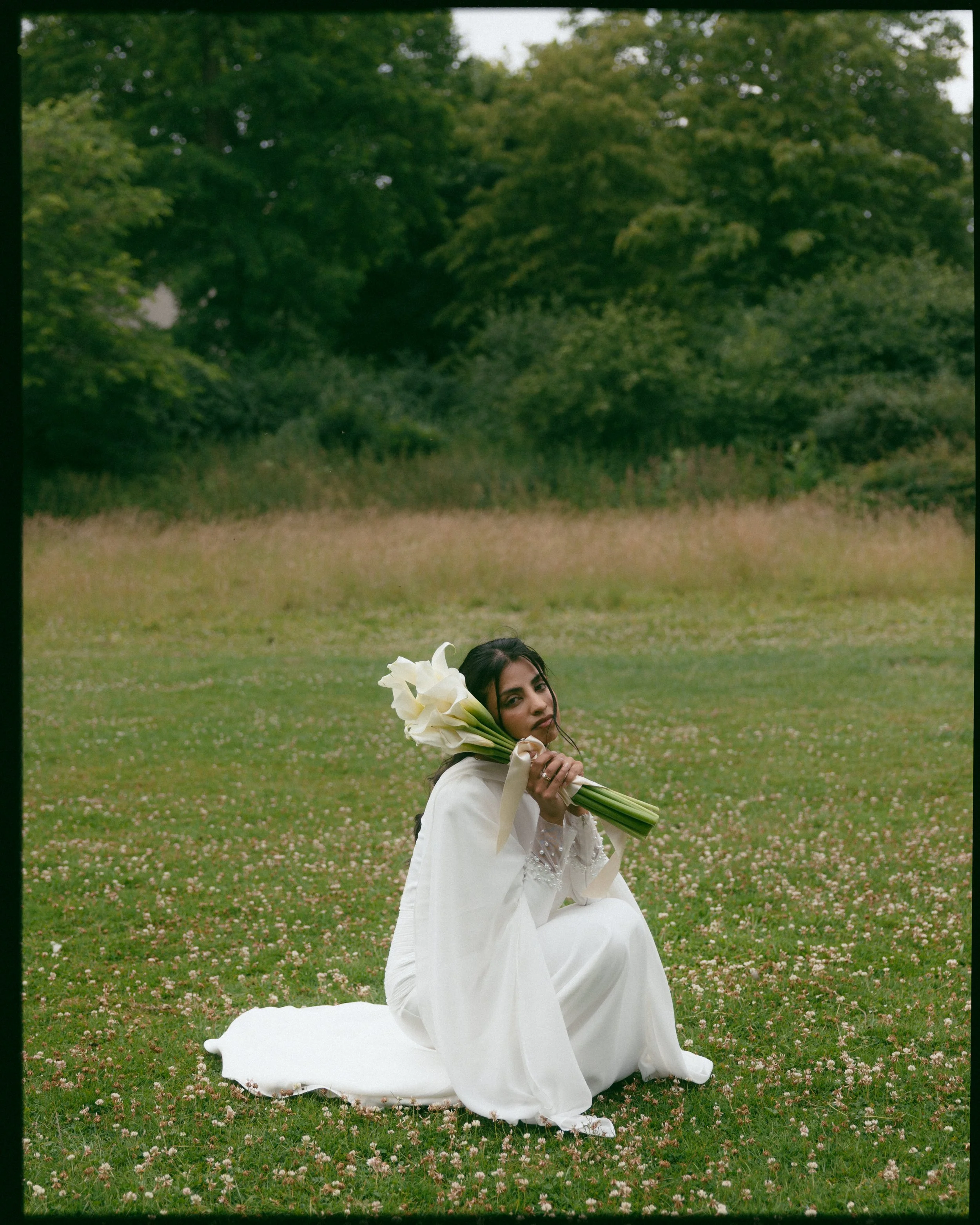 A woman dressed in white, sitting on the ground in a field of small pink and white flowers, holding a bouquet of large white calla lilies. She is outdoors with a background of green trees and grass.